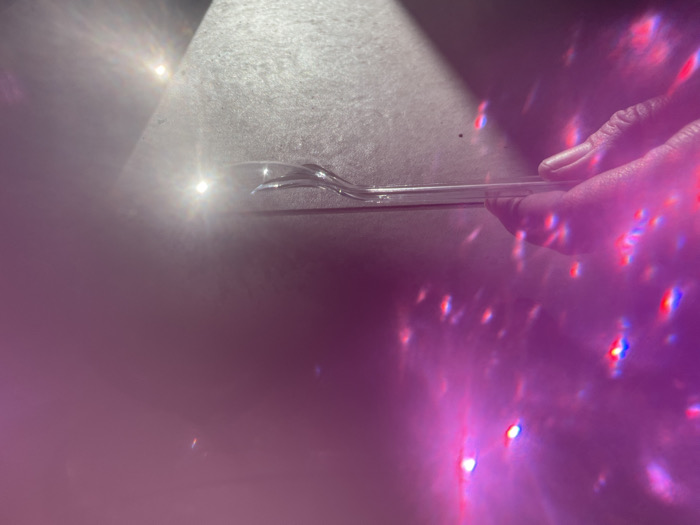 The scene depicts an indoor environment in Tel-Aviv, Israel, characterized by prominent light effects. A human hand, exhibiting light skin pigmentation and unpainted fingernails, is visible on the right side of the frame, holding a transparent utensil, possibly plastic, resembling a spoon or spork. The utensil's bowl is positioned towards the upper left, and its handle extends horizontally. This utensil appears to be slightly elevated above or resting on a light-colored, textured surface. A strong, localized light source reflects brightly off this surface in the upper left quadrant. The right and lower portions of the image are heavily overlaid with numerous scattered light reflections and flares, predominantly displaying red, blue, and pink/purple chromatic components.
