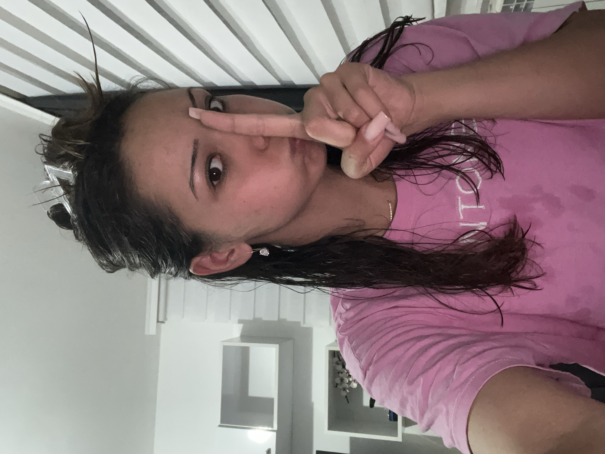 A young adult female is depicted in an indoor setting in Zlatograd, Bulgaria, captured in a mid-shot. Her right index finger (viewer's left) is positioned vertically over her lips, partially obscuring her face, while her cheeks are puffed out, creating a playful expression. Her left eye (viewer's right) is open and looking directly at the camera. She has dark, wet hair, pulled back with several clear hair clips visible at the crown, and several wet strands hang loosely around her face and shoulders. She wears a pink short-sleeved t-shirt with partial white lettering visible on the front, a delicate gold-toned necklace, and a single silver-toned drop earring with a clear stone on her right ear (viewer's left). Her fingernails are long and manicured in a light pink or nude color. The background features white walls. To the viewer's left, three white, rectangular floating shelves are mounted on the wall; the lowest shelf displays decorative items, possibly artificial flowers. To the viewer's right, vertical white window blinds are visible. The scene is illuminated by bright, artificial light.