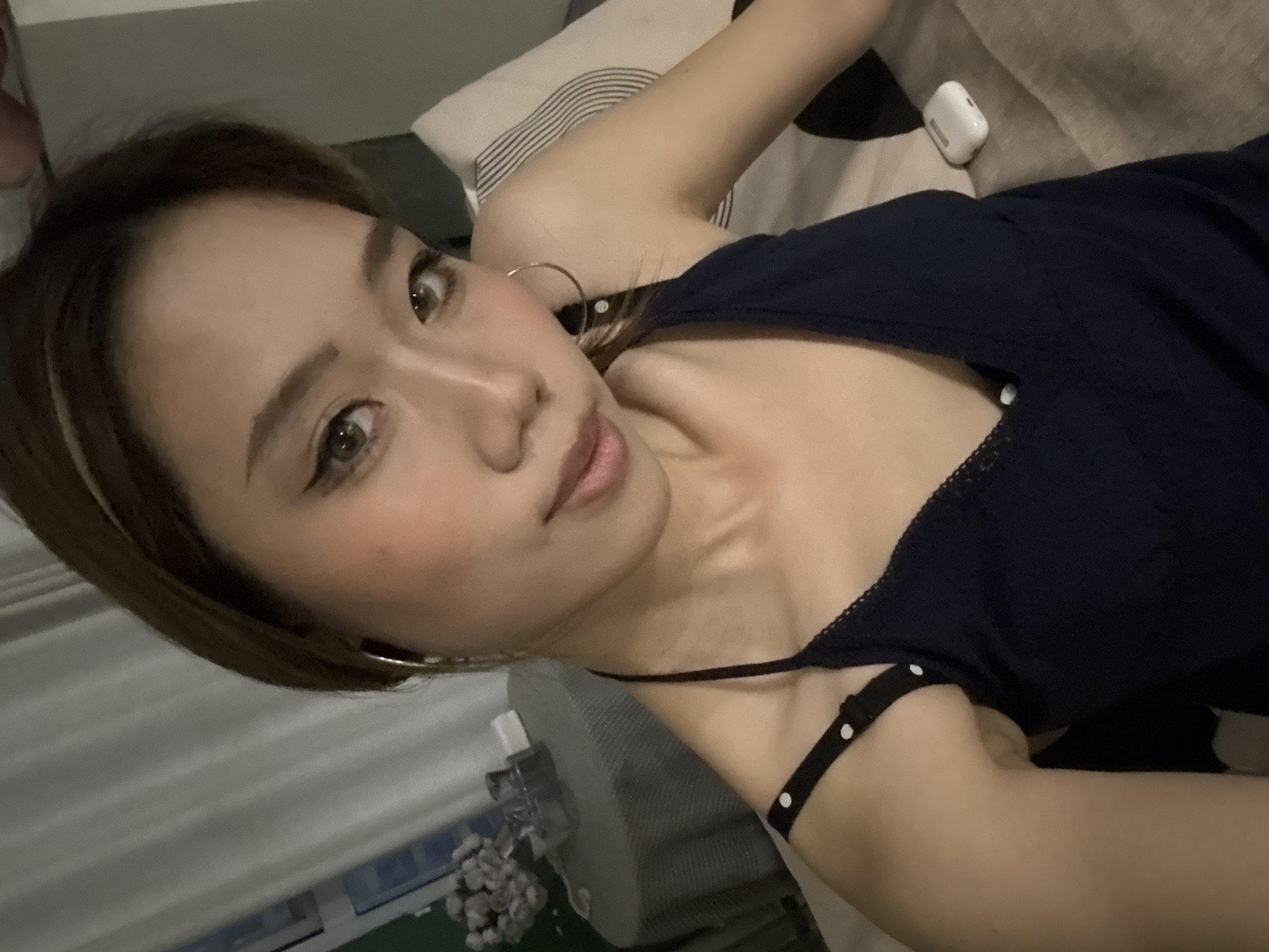 A woman with brown hair and fair skin is lying on a bed. She is wearing a dark blue camisole with white polka dots on the strap. She has a pair of gold hoop earrings. A white pair of earbuds are on the bed beside her arm. White curtains can be seen in the background. The location is not discernible from the image.