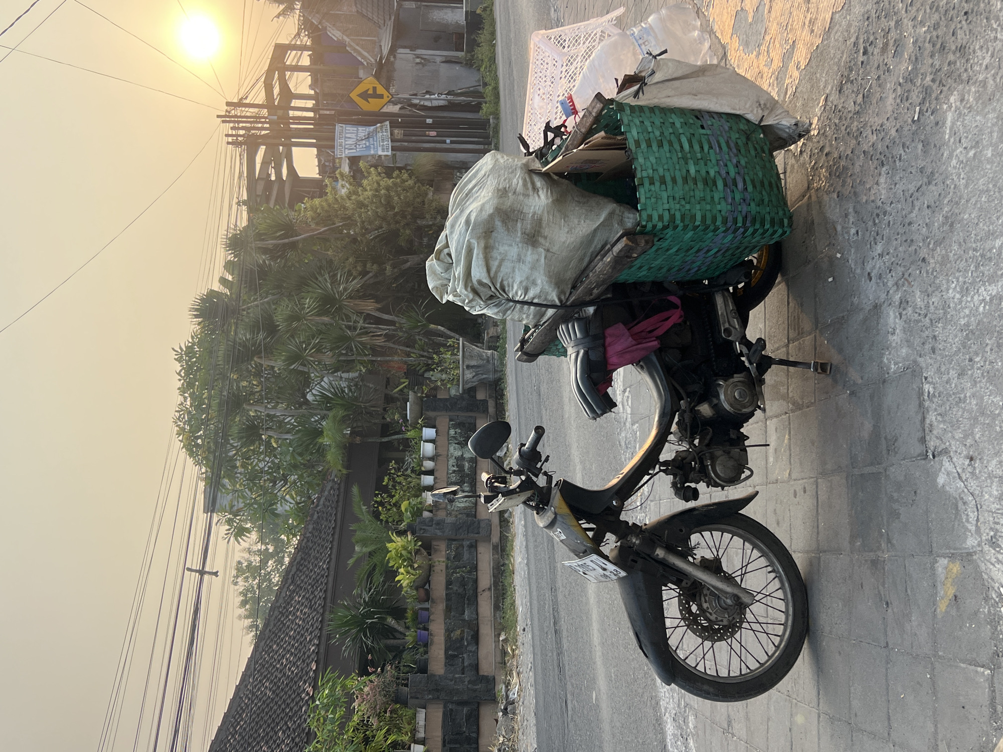 The image captures a weathered, black motorcycle loaded with various items, parked on a sidewalk in Tlogoadi, Indonesia. The motorcycle is a scooter-style model, with a license plate visible. A large, tan-colored sack is prominently secured to the rear of the motorcycle, along with a green woven basket overflowing with what appears to be recyclables, including plastic bottles and cardboard. The motorcycle is situated on a gray, paved sidewalk adjacent to a street. In the background, houses with red-tiled roofs and lush green vegetation are visible. Utility wires crisscross the sky, and the setting sun casts a warm, hazy glow over the scene, suggesting it is late afternoon or early evening. A yellow directional sign with an arrow is affixed to a pole in the middle ground, and there is evidence of ongoing construction in the distance. The overall impression is a snapshot of daily life in a residential area of Tlogoadi, with the motorcycle likely used for collecting or transporting goods.