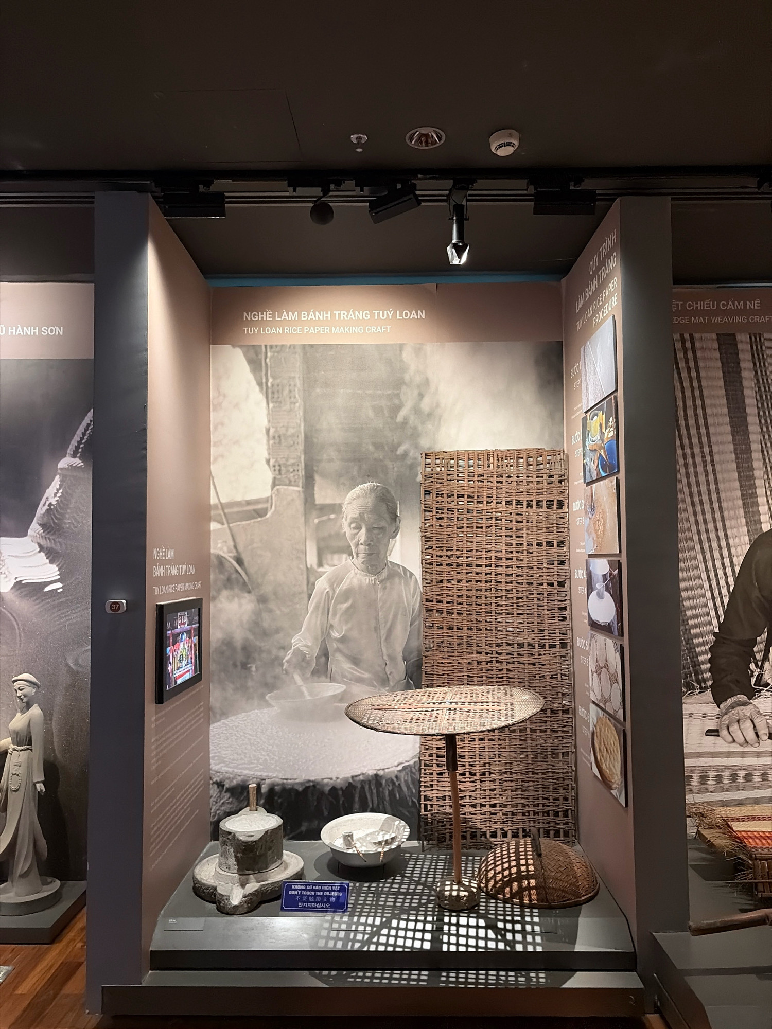 The image shows an exhibition display for "Tuy Loan Rice Paper Making Craft" in Da Nang, Vietnam. The display features a large black and white photograph of an elderly Vietnamese woman making rice paper, surrounded by artifacts related to the craft. These artifacts include a stone mill, a bowl, a woven mat on a stand, and a woven basket. Text panels in Vietnamese and English provide information about the craft. To the left of the main display, a statue of a woman in traditional attire is visible. The exhibition appears to be located in a museum or cultural center.