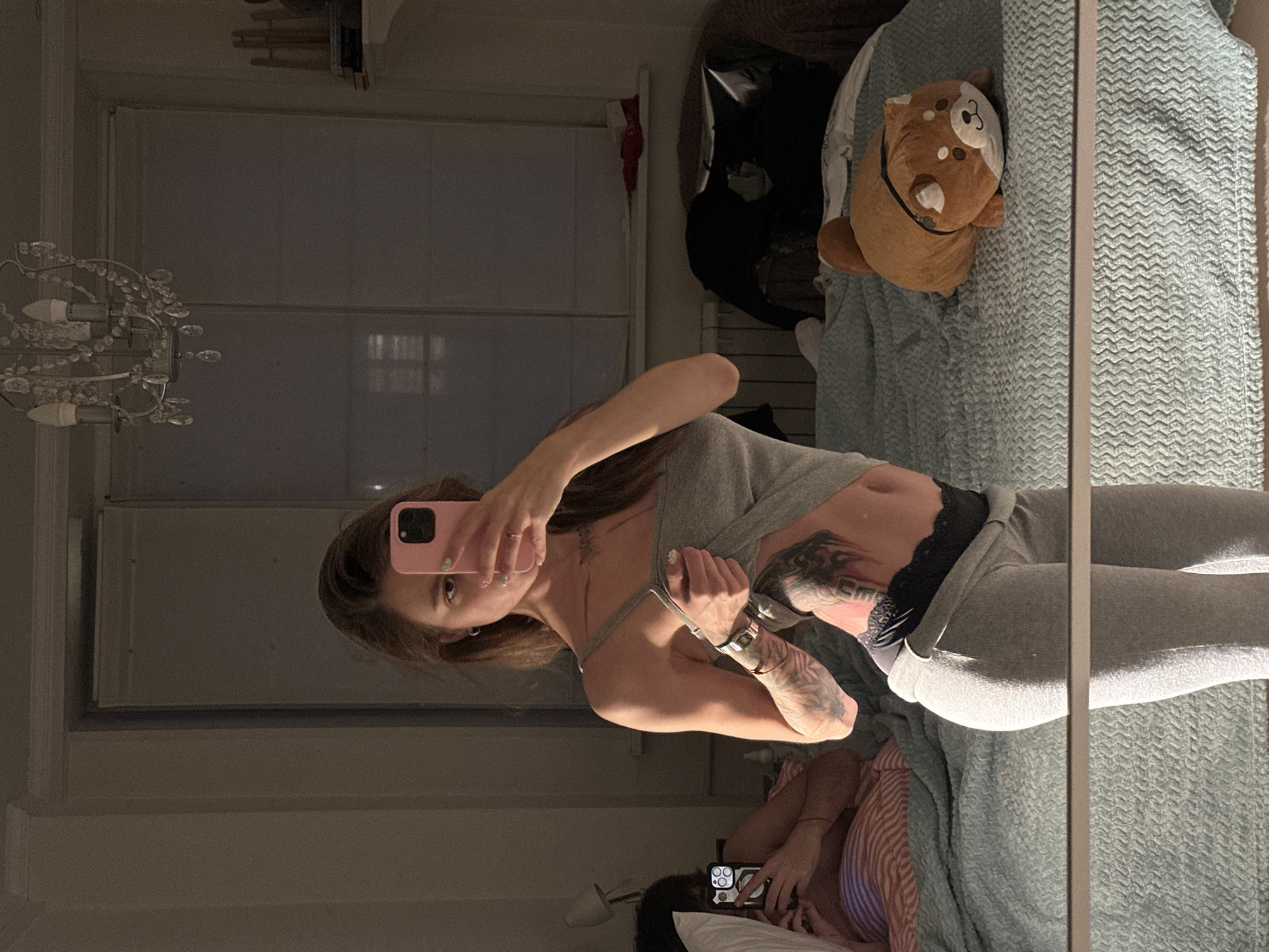 A woman is taking a mirror selfie in a bedroom. She is wearing a grey tank top, black lace underwear, and grey sweatpants. She has tattoos on her torso and arm. The room has a bed with a plush dog toy on it, a dresser with books, and a chandelier.