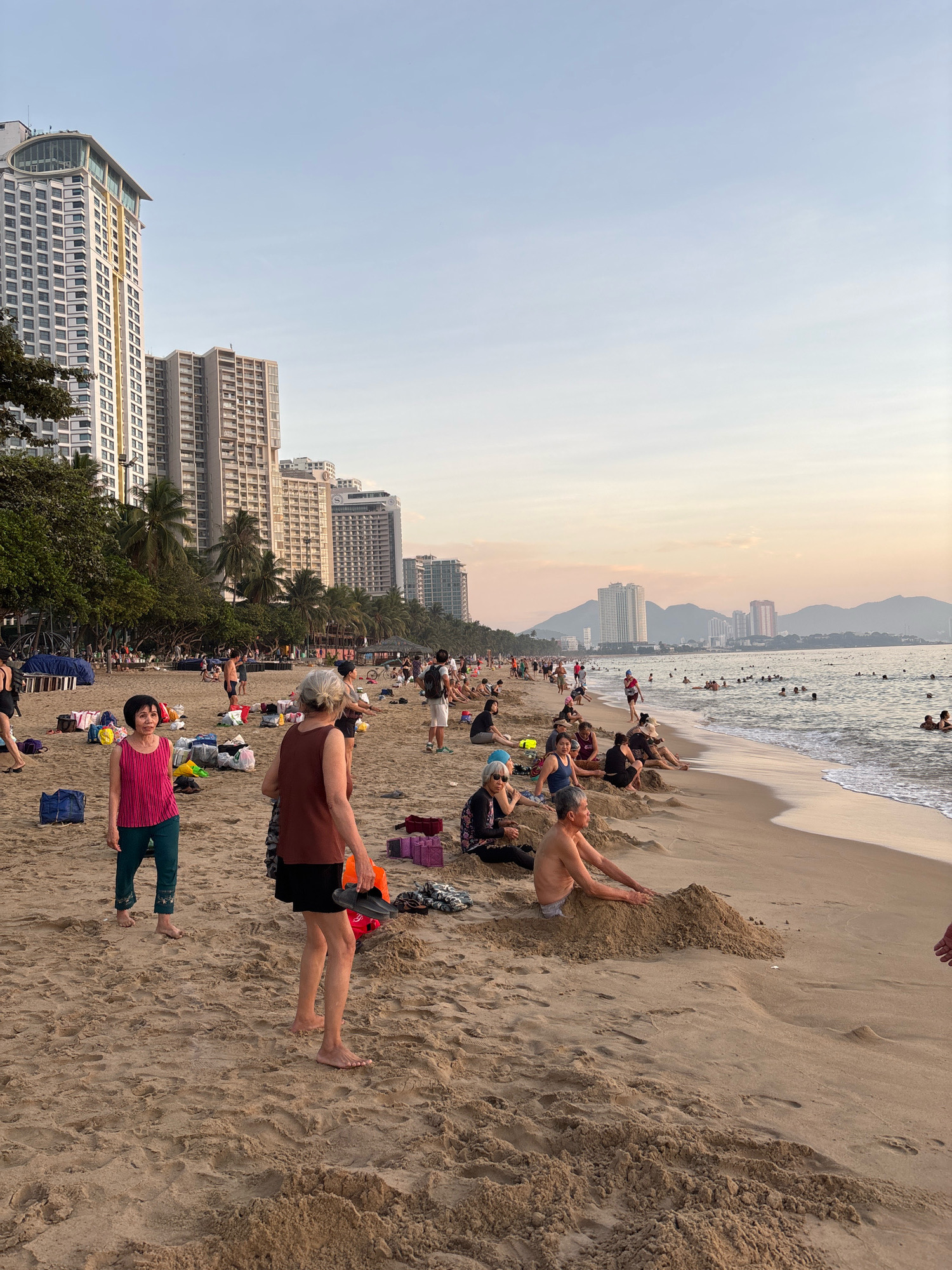 The image depicts a crowded beach scene in Nha Trang, Vietnam, during the late afternoon or early evening. Tall residential buildings line the coastline, with lush green trees and palm trees interspersed between them. The beach itself is sandy, with numerous people engaged in various activities.
In the foreground, two women are standing on the sand. One woman, wearing a pink sleeveless top and green pants, stands facing left. The other woman, in a brown sleeveless top and black shorts, stands facing right, holding a pair of dark sandals in her right hand. Several beach bags and other items are scattered on the sand around them.
Further down the beach, a man is partially buried in a mound of sand, with his arms resting on the sand pile. Other people are seen sitting and lounging on the sand, some in groups, while others are walking. In the background, the ocean is visible with many people swimming and enjoying the water. The sky is a clear pale blue, transitioning to a soft orange and pink hue near the horizon, suggesting the sun is setting. Distant mountains can be seen across the water. The overall atmosphere is one of leisure and recreation.