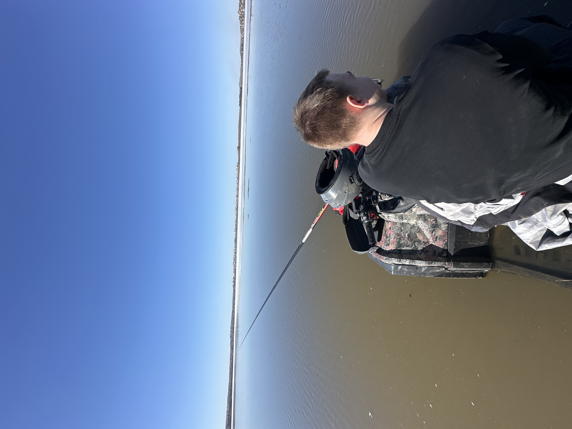 A man sits on a small watercraft in a body of water, possibly a lake or wide river, on a clear day with a cloudless blue sky. The shore is visible in the distance on the left side of the image. The man is wearing a black long-sleeved shirt. The watercraft appears to be camouflaged and has a helmet attached to it. A long pole or fishing rod extends out into the water. The location is Unknown, Unknown.