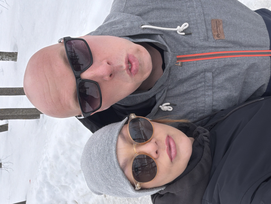 Two individuals are depicted in a close-up, selfie-style photograph outdoors within the city of Chaikovsky, Russia, against a snowy background. The person on the left is a female, wearing a grey knit beanie, round light-colored translucent frame sunglasses, and a dark, possibly black, winter jacket. Her lips are slightly parted, showing light pink lipstick. The person on the right is a bald male, wearing black-framed sunglasses and a grey winter jacket with an orange main zipper and white hood drawstrings. A brown brand patch, visible on his right chest area, reads "OUTVENTURE". His mouth is open in an O-shape. Both individuals are looking directly at the camera. The background consists of a snow-covered ground and several bare deciduous trees with light-colored bark.