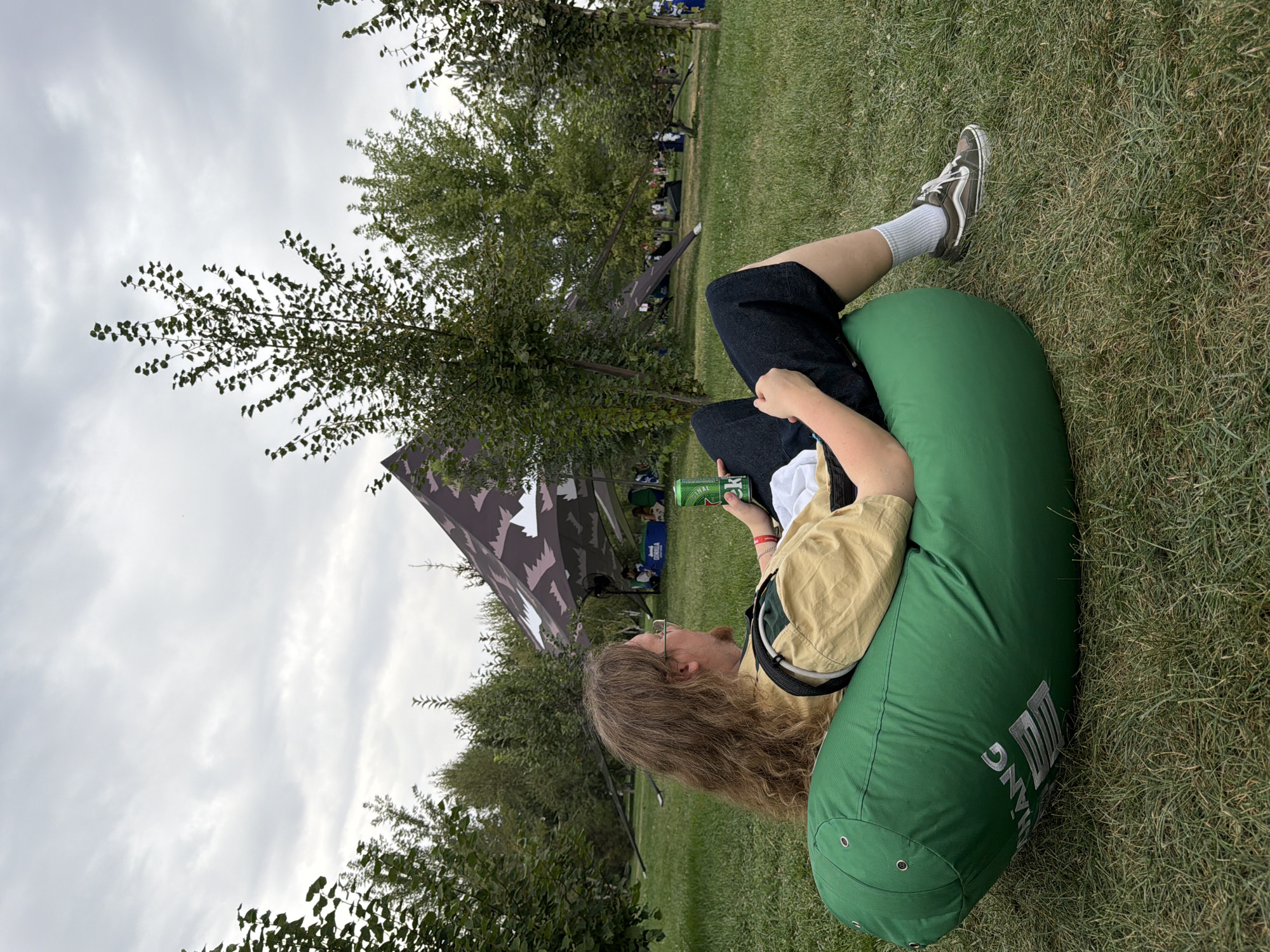 In Pervomayskiye Prudy, Kazakhstan, a person with long, wavy brown hair and glasses sits reclined on a large, green beanbag chair on a grassy lawn. They are wearing a light yellow shirt with dark trim and dark shorts, and are holding a green can. The background features several trees, a large patterned tent structure, and glimpses of other people in the distance. The sky is overcast.