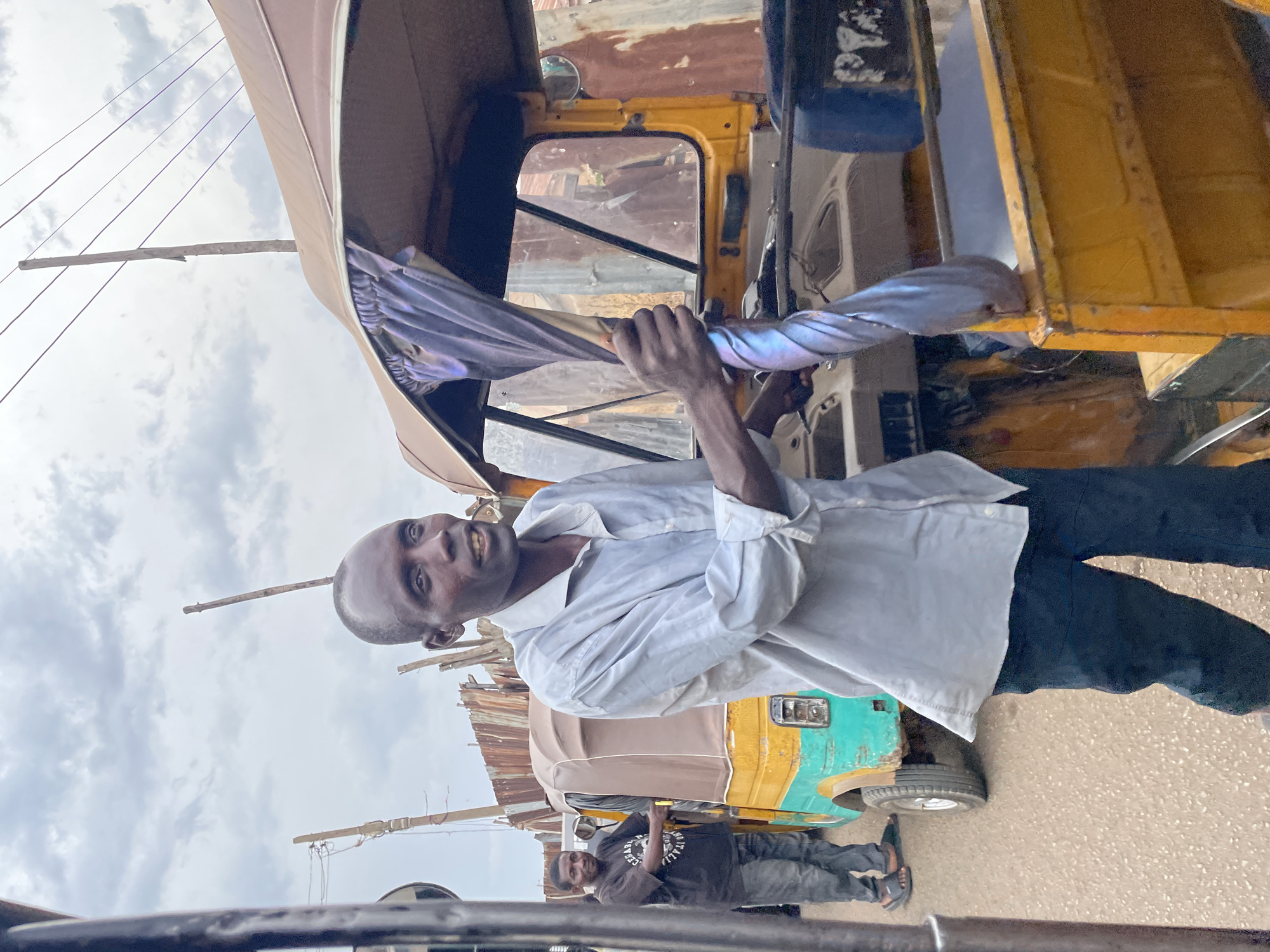 The image captures an outdoor scene in Jos, Nigeria, featuring two men and tricycles under an overcast sky.
In the foreground, a dark-skinned man wearing a light blue collared shirt and dark trousers stands looking directly at the viewer with a slight smile. His left arm is bent, and he holds onto a twisted, shiny silver-colored fabric or material that appears to be part of a vehicle or a decorative element. He is positioned next to a light brown and beige-colored tricycle (locally known as a keke napep), which is partially visible behind him.
Further in the background, to the right, another dark-skinned man stands casually. He is wearing a black t-shirt with the text "MADE IN ITALY" clearly visible across the chest, and dark jeans. Behind him, a green and yellow tricycle is parked, and a backdrop of corrugated metal structures suggests an urban street or market area. Overhead, several utility wires are strung between wooden poles against a cloudy, bright sky.
The activity depicted is casual standing or posing next to vehicles in what appears to be a typical daytime street environment. The weather is bright but overcast, with no direct sunlight visible. The scene reflects an everyday moment in a Nigerian city, with local transportation (tricycles) prominent.