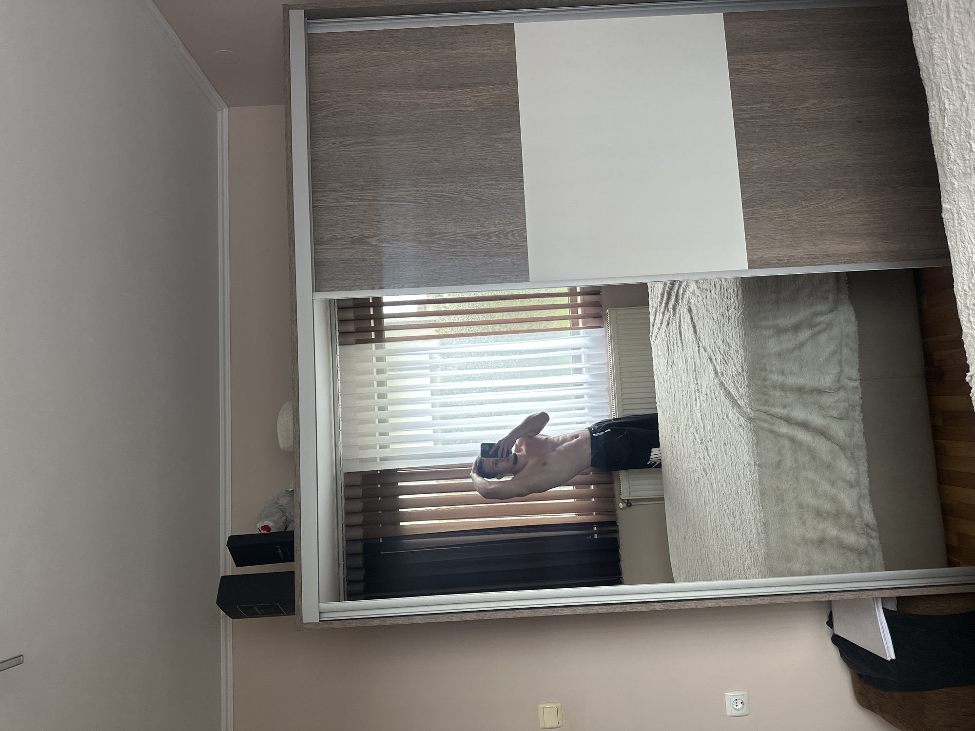 This image depicts an indoor scene, specifically a bedroom, located in Saraia, Bulgaria. The central focus is a full-length mirror, which is a sliding door of a wardrobe, reflecting a male individual and portions of the room.
The person reflected in the mirror is an adult male, shirtless, with a muscular physique, displaying visible abdominal muscles. He is holding a black smartphone with his right hand, positioned to capture his reflection. His left arm is raised, with his hand placed behind his head. He is wearing black athletic shorts that feature a white logo on the left leg, consistent with the "Nike" brand. The individual's facial features are visible through the reflection in the phone screen.
The wardrobe, where the mirror is integrated, consists of multiple sliding doors. The mirror occupies one entire door. Adjacent doors exhibit a two-tone design: an upper section with a greyish-brown wood-grain pattern and a lower section in plain white.
The room reflected in the mirror features light peach-colored walls. A large window dominates the background, fitted with vertical blinds; some are dark brown and opaque, while others are sheer white, allowing diffused light and a blurred view of green foliage outside. Below the window, a white horizontal radiator is mounted. The room's ceiling is white, adorned with a decorative white cornice molding along its perimeter. A large bed is visible, covered with a textured, fluffy, off-white or light grey comforter. The bed frame appears to be upholstered in a light beige fabric. A segment of wooden flooring is visible to the right of the bed. On the left wall, a light-colored rectangular light switch panel is present, along with a white electrical double socket positioned lower on the wall. Adjacent to the socket, a dark piece of clothing and a white notebook or book are resting on a brown upholstered surface, possibly a chair or bench.
On top of the wardrobe, outside the direct reflection of the room, several objects are visible: two dark, rectangular boxes, one bearing the text "CHANEL" and the other "INSOMNIA"; a small, grey, fluffy object; and a light-colored, possibly woven, circular item.
The scene is illuminated by natural daylight entering through the window.