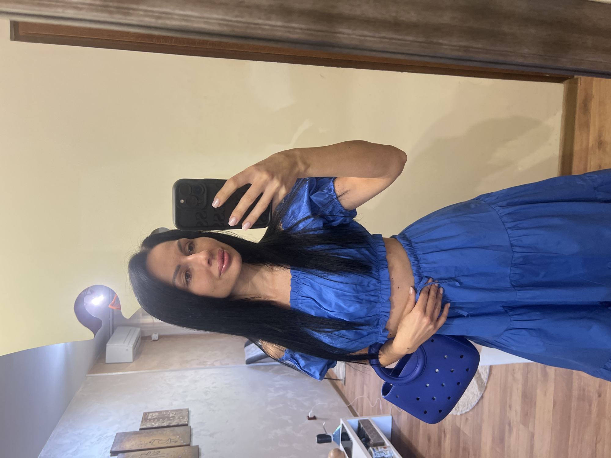 A woman is taking a mirror selfie in a room. She has long, dark hair and is wearing a blue off-the-shoulder crop top and a matching tiered maxi skirt. She is holding a blue perforated tote bag in her left hand. The room appears to be a home, with a wall mounted air conditioning unit visible in the background, along with artwork on the wall. The floor is wooden. The location is in the city of Stara Zagora, Bulgaria.