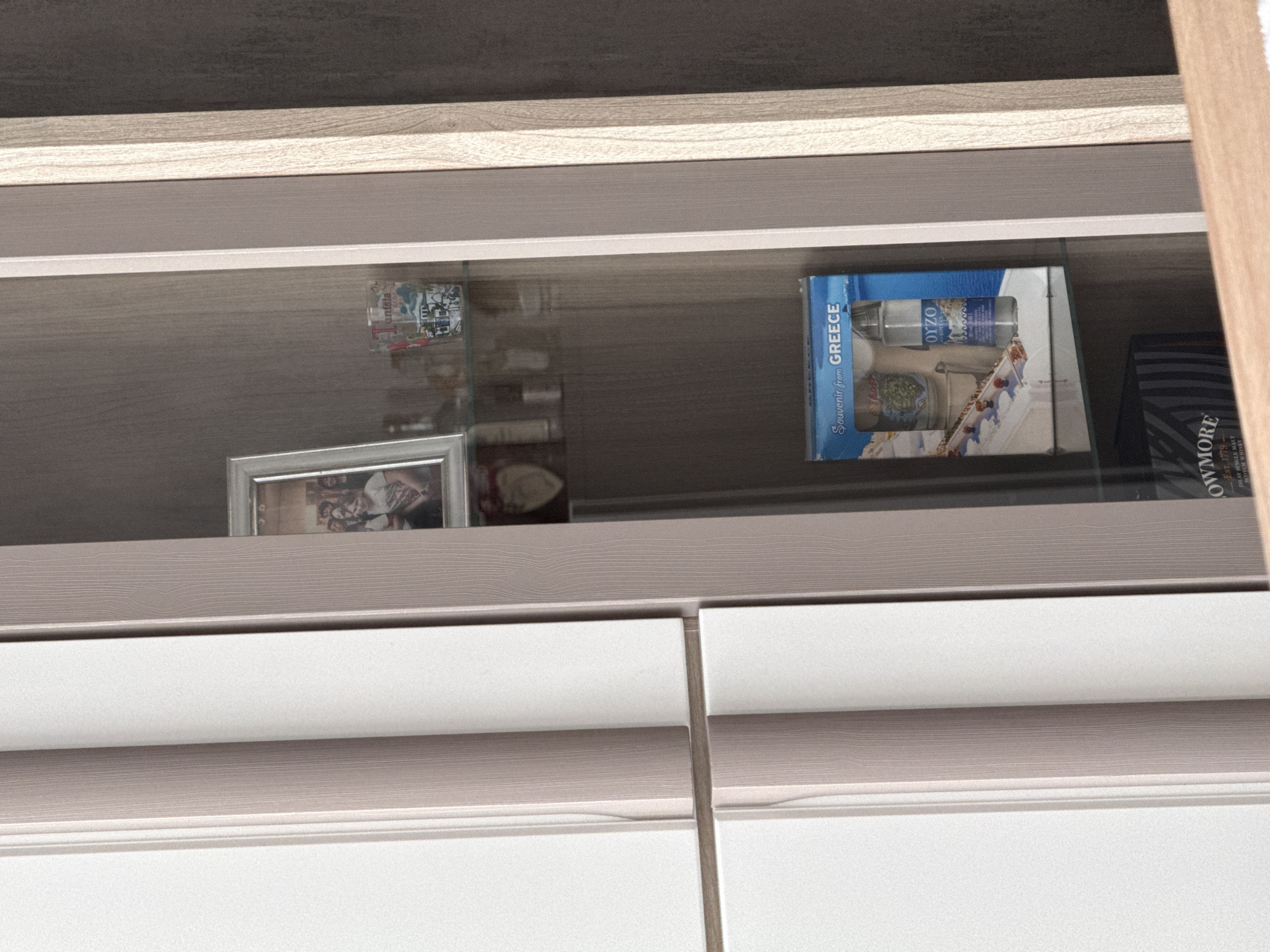 The image depicts a section of a cabinet or shelf, likely in an interior space. The shelf contains various items, including a souvenir box from Greece, a framed photograph, and other smaller objects that appear to be decorative. The cabinet doors below the shelf are white with a horizontal band of light wood-grain texture. The overall environment suggests a modern or contemporary home setting. There is no evidence to suggest it is in Lovech, Bulgaria.