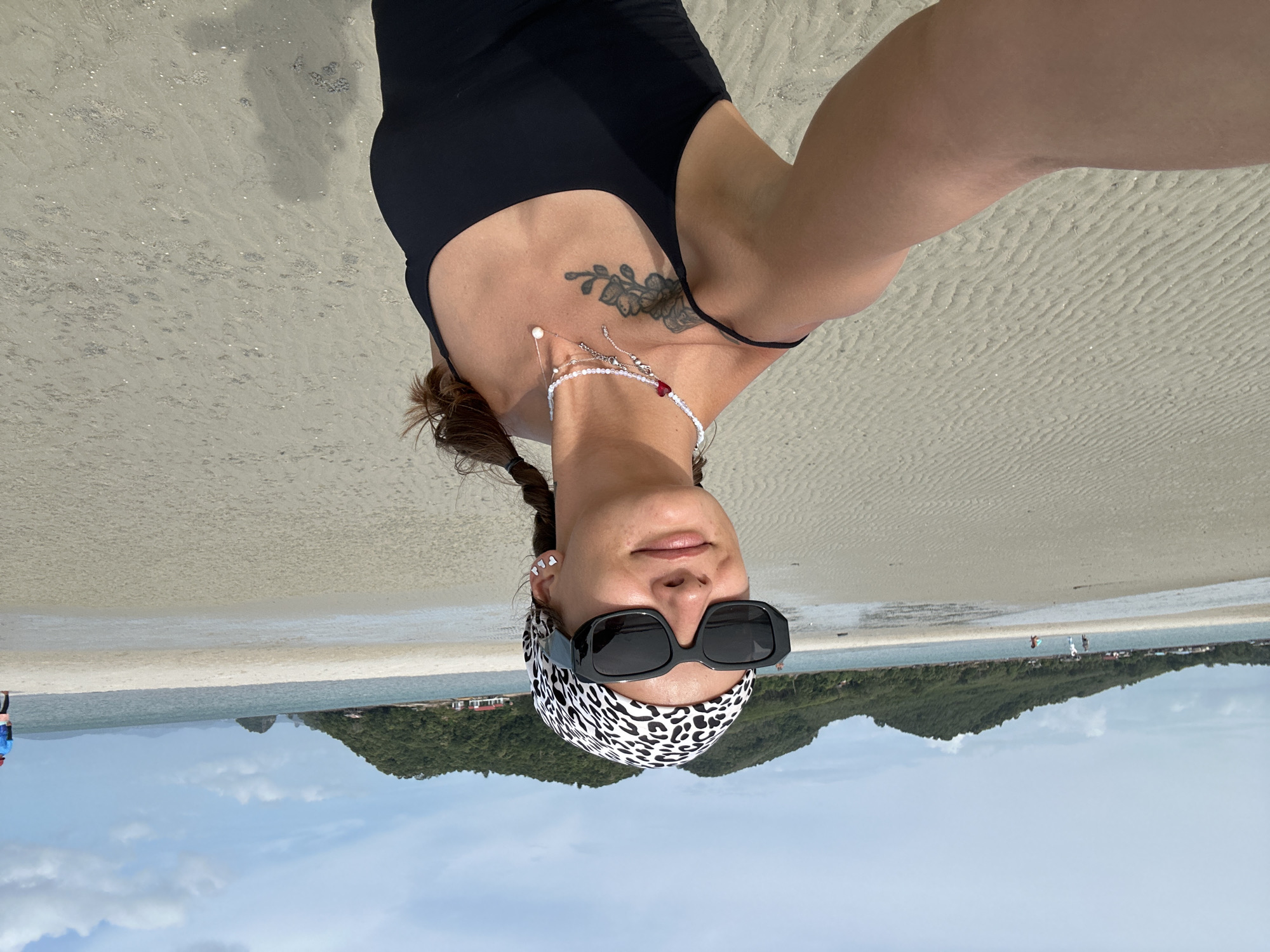 A woman is taking a selfie on a sandy beach. She is wearing a black tank top, a leopard print headscarf, and large black sunglasses. She has a tattoo on her left shoulder and is wearing several necklaces. Her hair is in two braids. The beach is wide and flat, with ripples in the sand from the receding tide. In the background, there is a calm body of water, a distant green island with trees, and a cloudy sky. A few other people are visible in the distance, walking near the water's edge. The scene appears to be set in Ko Yao, Thailand.