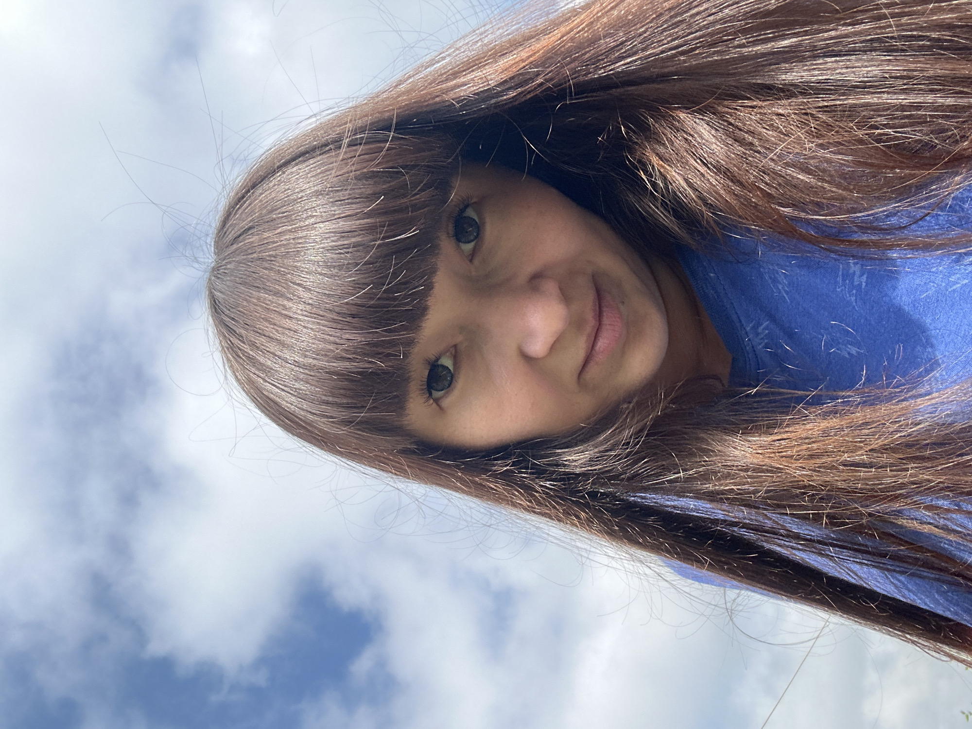 A medium shot captures a person looking up at the camera against a backdrop of blue sky with white clouds. The person, a woman with long, brown hair and bangs, is wearing a blue t-shirt. Her face is framed by her hair, and the camera angle is from below, looking upwards. The lighting suggests it is daytime. The specific location in the city of Sredneye Kechevo, Russia, is not discernible from the visual information provided in the image.