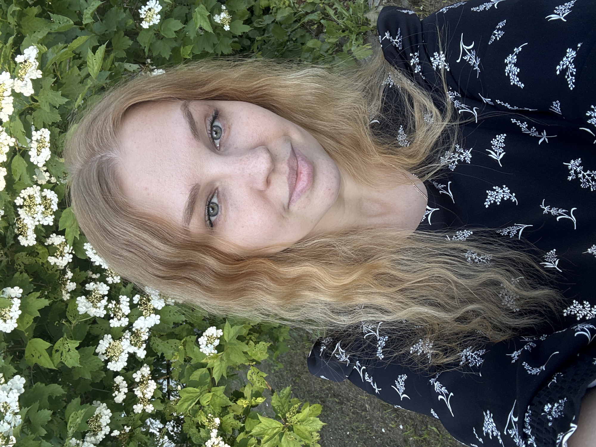 A young adult female is depicted in a close-up outdoor portrait in Lepilovo, Russia. She has long, wavy, light blonde hair parted down the center, green eyes, and visible freckles on her nose and cheeks. Her lips are slightly upturned in a gentle smile, and she is looking directly into the camera. She wears a black garment, likely a dress, adorned with a repeating pattern of white stylized floral or leaf motifs. A thin, gold-toned chain necklace is visible on her left side. The background consists of a dense green bush with numerous clusters of small white flowers, each with yellowish centers. Some leaves on the bush show signs of insect damage. The lower left portion of the image reveals sparse ground cover with grass and soil.