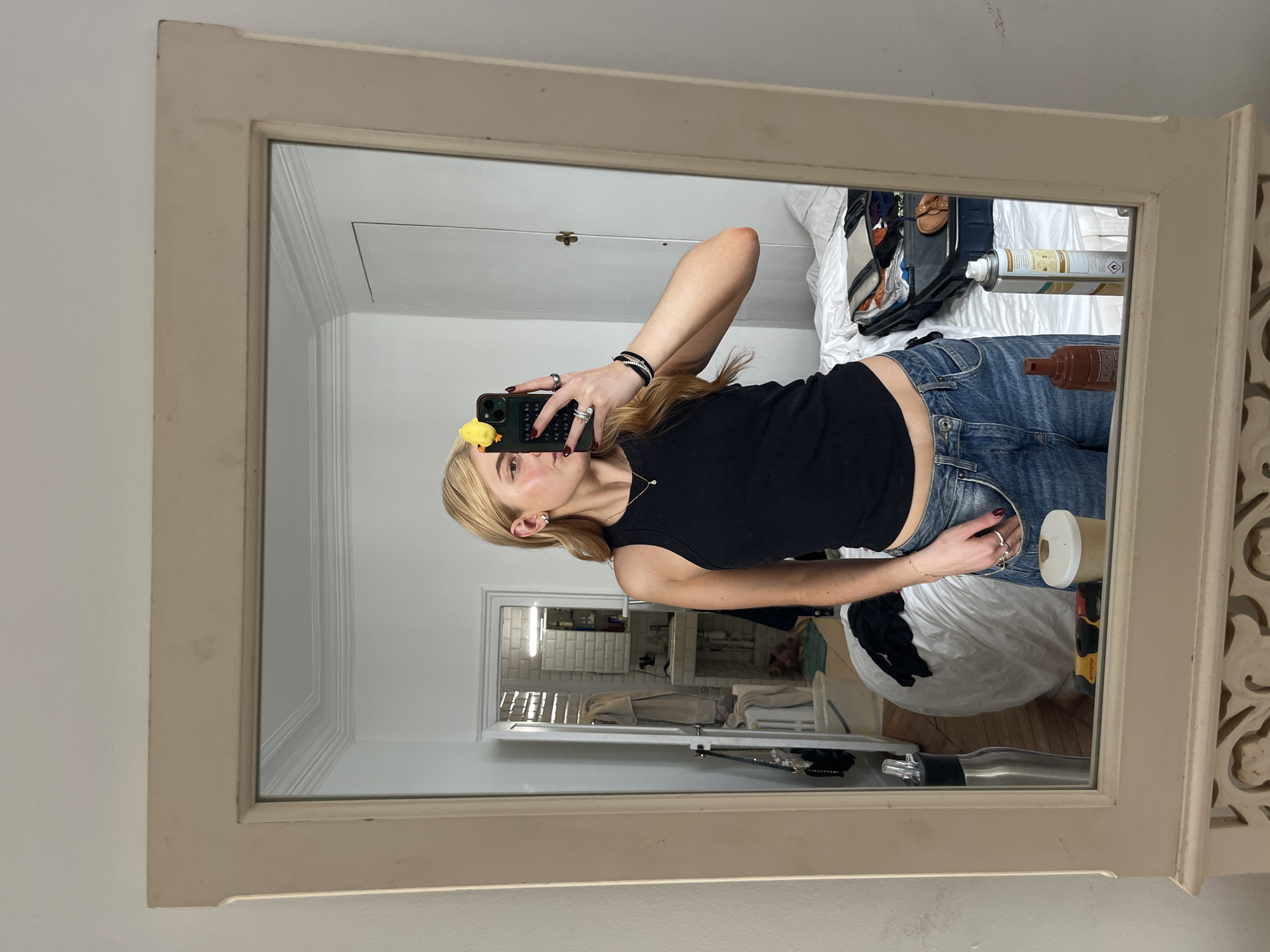 A person stands in front of a mirror taking a selfie. The person is wearing a black tank top and blue jeans. The person is holding a phone with a yellow object attached to it. The background shows a room with a bed, a white blanket, and various items scattered around. Through an open doorway in the mirror reflection, a tiled bathroom can be seen. The location of the photo is most likely not in Paris, France.
