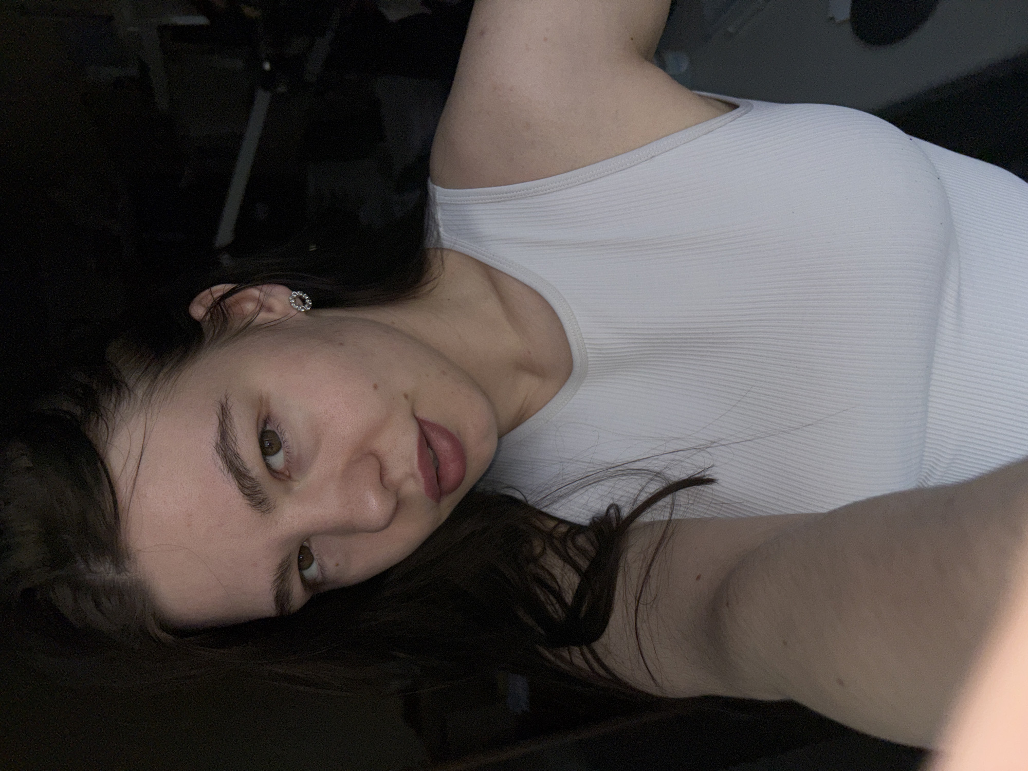 A woman with fair skin and dark brown hair is lying down, her head tilted to the left, with the lower right portion of her face facing the camera. She has brown eyes, and her lips are slightly parted. She is wearing a ribbed, light-colored tank top. A small, round earring is visible in her left ear. The background is dark and indistinct, but a tripod or other equipment can be partially seen behind her head. The location in the city of Perm, Russia, is not discernible from the image.