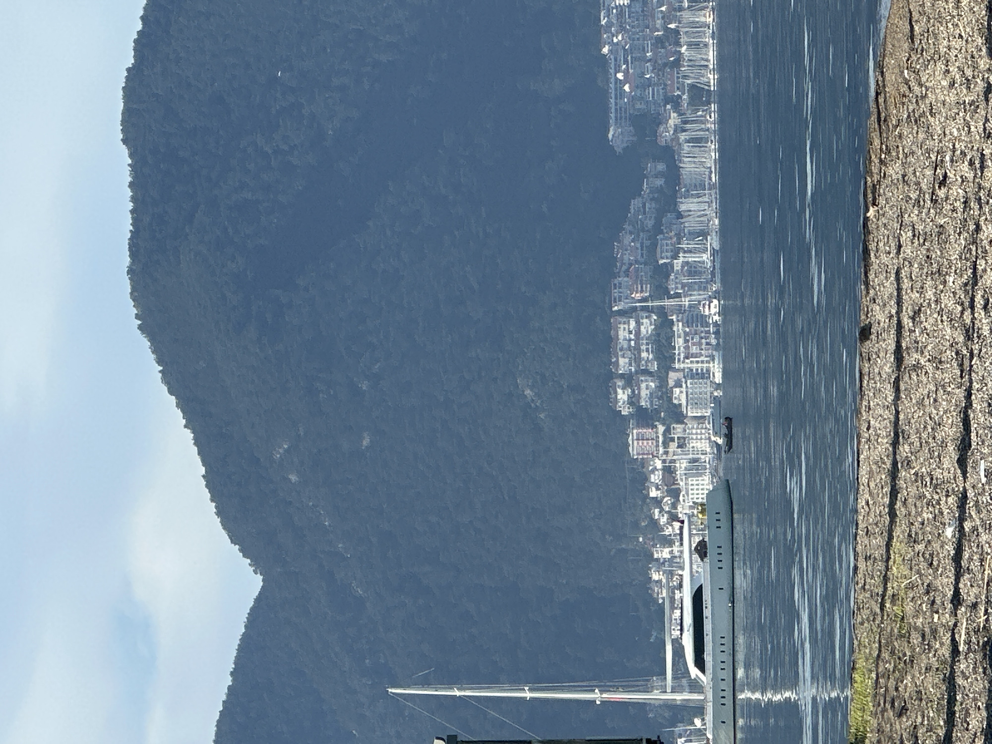 The image shows a landscape in Fethiye, Turkey, featuring a body of water (likely the Aegean Sea) with a large yacht positioned on it. The yacht appears to be a modern sailing vessel. Along the shoreline opposite the yacht, there's a dense collection of buildings, suggesting a populated area within Fethiye. A large, green mountain looms in the background, partially obscured by the buildings and water, further defining the area's topography. In the immediate foreground on the right is a retaining wall.