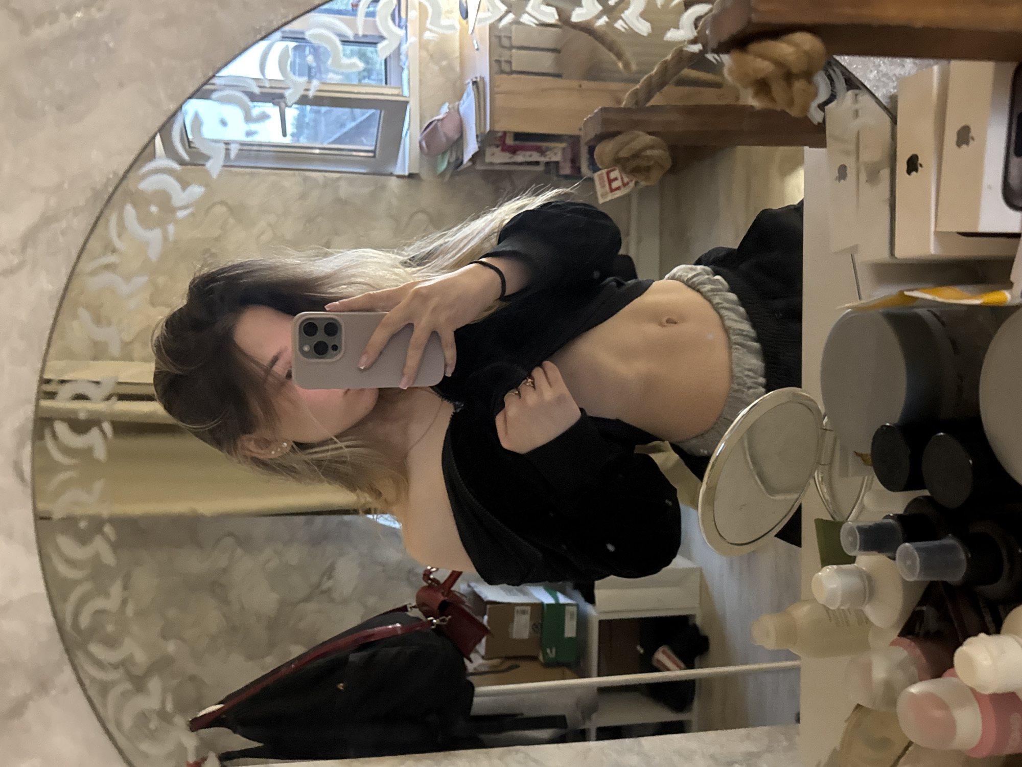 A young woman is taking a selfie in a mirror. She is wearing a black sweater and grey pants. Her stomach is showing. The background includes a window, a cat tree, and a shelf with various items on it. There is no clear evidence of the location of Noviy, Russia in the image.