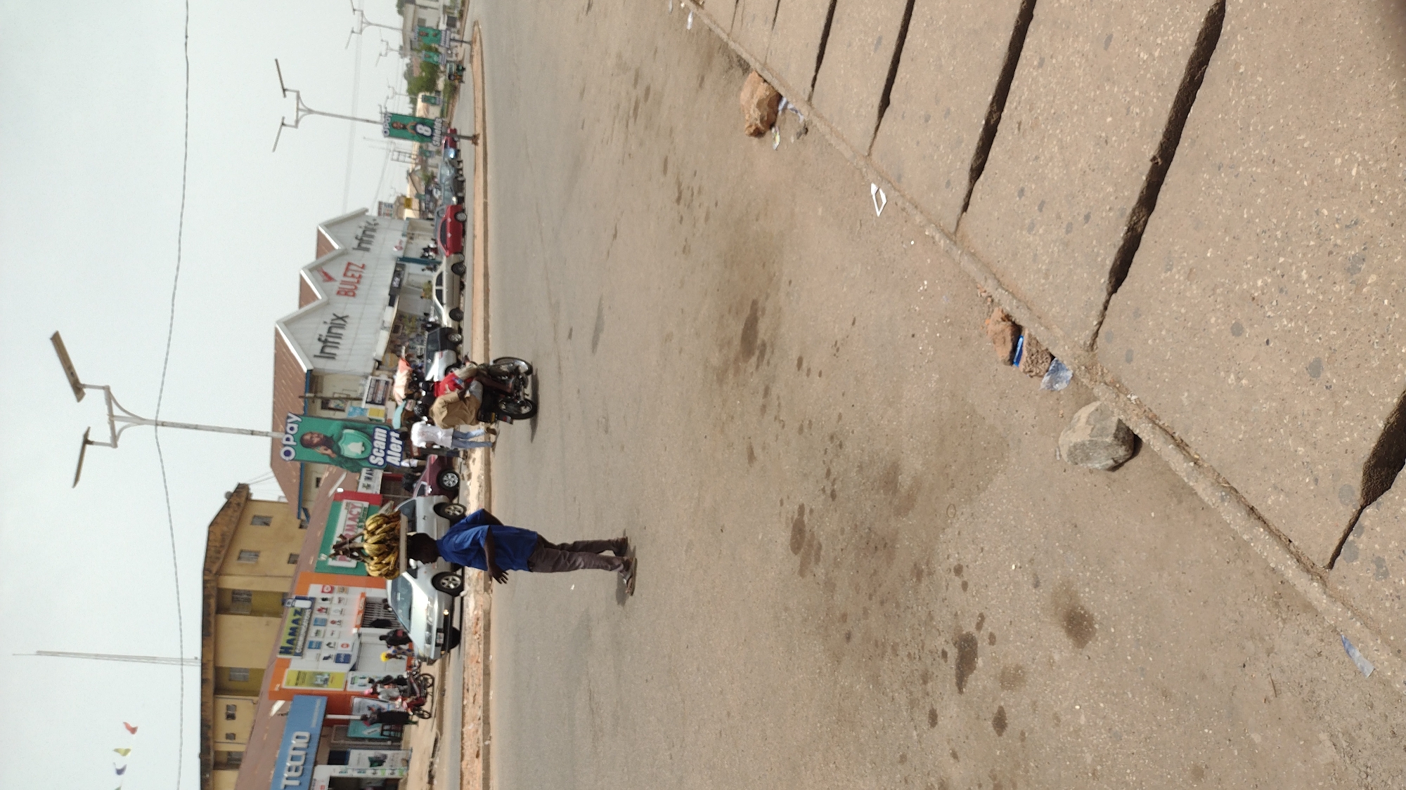 The image captures a street scene in Jos, Nigeria. A man in a blue shirt and dark pants stands on the asphalt road. In the background, there are several stores and buildings, including an Infinix and Buletz store. Cars and motorcycles are parked along the roadside. The street is relatively empty, with a few people visible. There are some rocks scattered near the curb of the road.