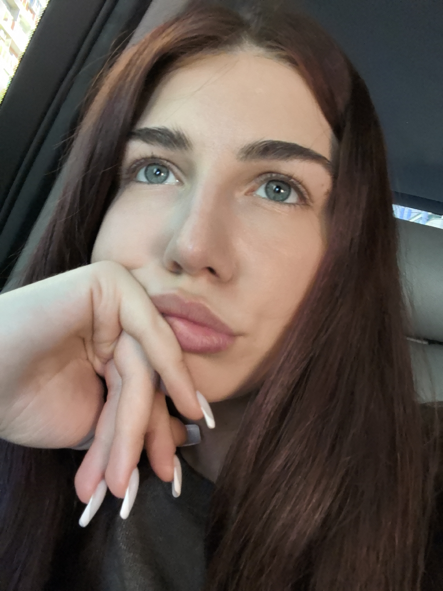 A young woman with long, dark brown hair and blue eyes is captured in a close-up, high-angle selfie, likely taken inside a vehicle. She rests her chin on her hand, her fingers adorned with long, white, almond-shaped acrylic nails. Her expression is pensive, with her gaze directed upwards and slightly to the left. The background is out of focus, suggesting the interior of a car with a dark headrest visible on the right. The scene offers no specific visual cues to pinpoint a location within Stara Zagora, Bulgaria.