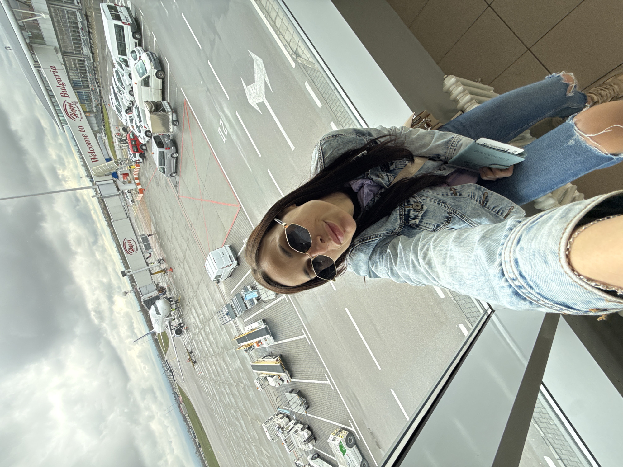 A medium eye-level shot shows a woman sitting inside an airport terminal in Sofia, Bulgaria. The subject wears a light blue denim jacket, ripped jeans, and sunglasses. She holds a passport and tickets. Outside the window is an airfield with various vehicles, including several white vans. A sign reads "Welcome to Sofia, Bulgaria." The weather appears overcast.