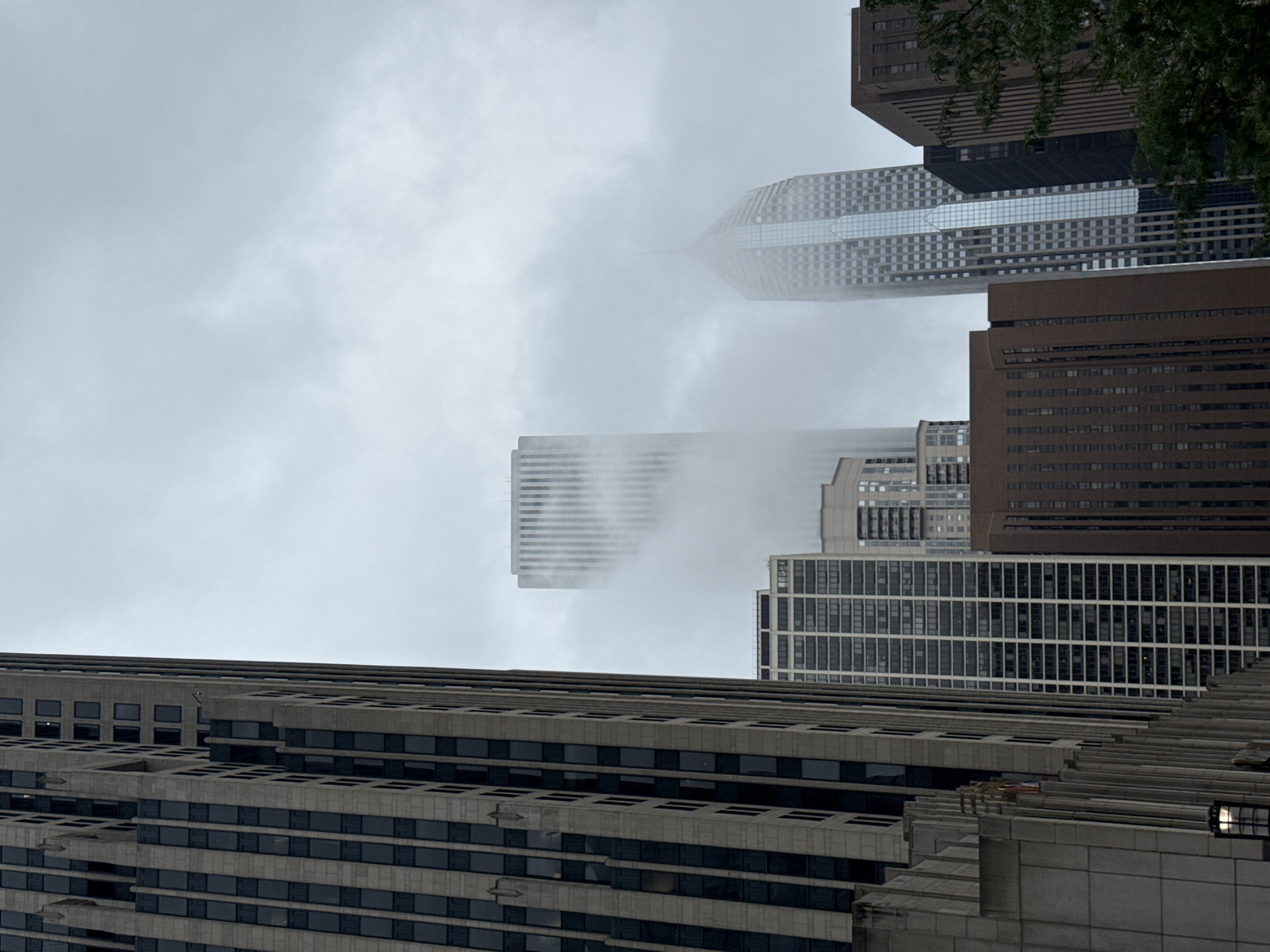 The image captures a cloudy day in downtown Chicago, Illinois, USA. The foreground is dominated by the imposing facade of a tall, art deco-style skyscraper. This building features a grid of dark windows set against a light-colored stone exterior, with vertical ribbed detailing and stepped setbacks. To the right of this building, other skyscrapers rise into the foggy sky. One of these is a sleek, modern tower with a glass facade that reflects the muted light, and another appears as a tall, slender structure partially obscured by mist. The lower portions of several other buildings are visible, displaying varying architectural styles and materials, including darker brick and concrete structures. The overall atmosphere is one of urban density under an overcast sky, with fog obscuring the upper reaches of some of the city's iconic architecture. No people or vehicles are visible in the frame.