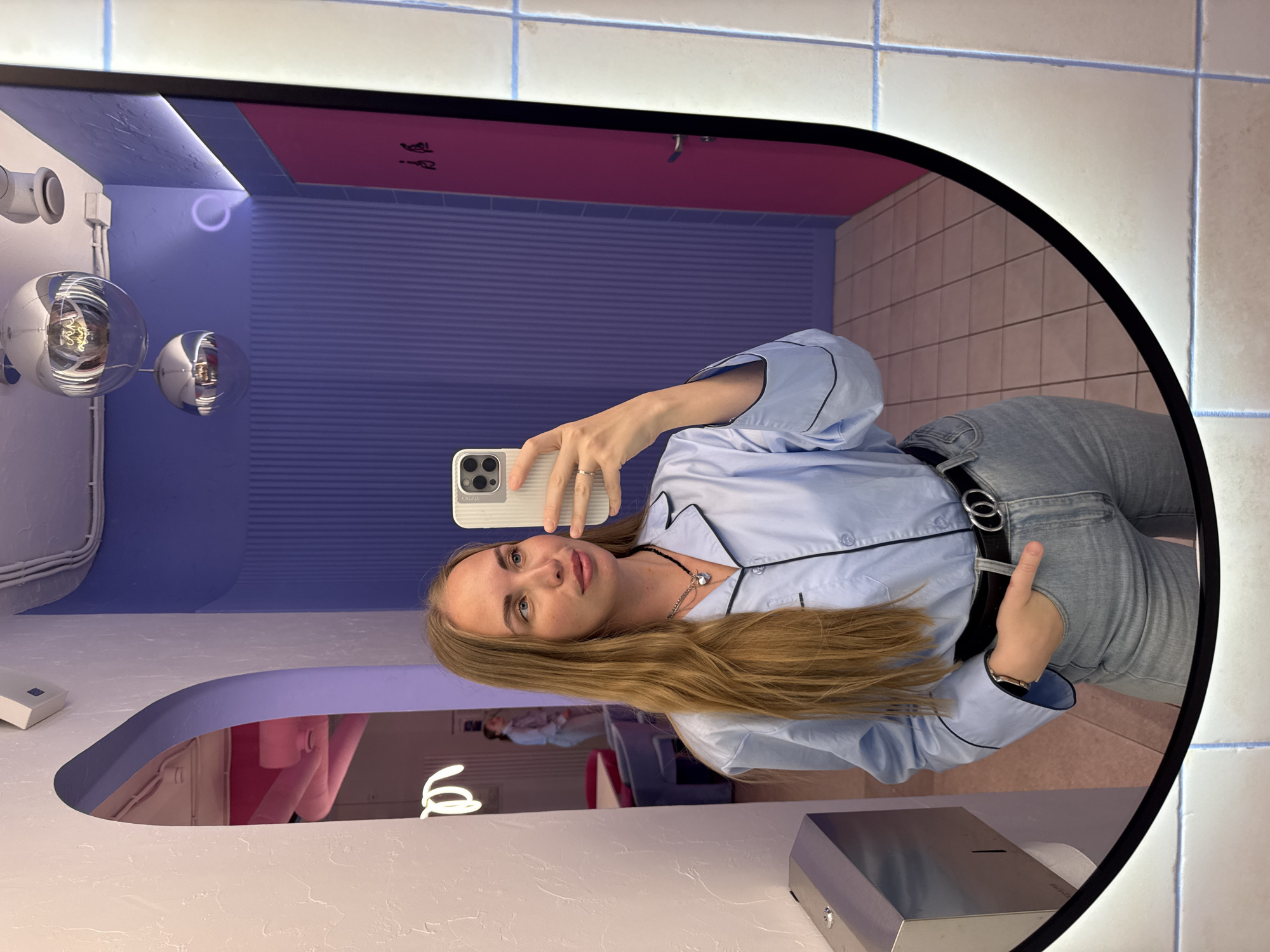 A young woman is taking a selfie in the mirror of what appears to be a public restroom. She is wearing a light blue pajama-style top with black trim and light wash jeans. The restroom features a modern design with a combination of purple and pink decor. The background includes a tiled wall and distinctive spherical light fixtures. Based on the provided information, the location is a restroom in the city of Sochi, Russia.