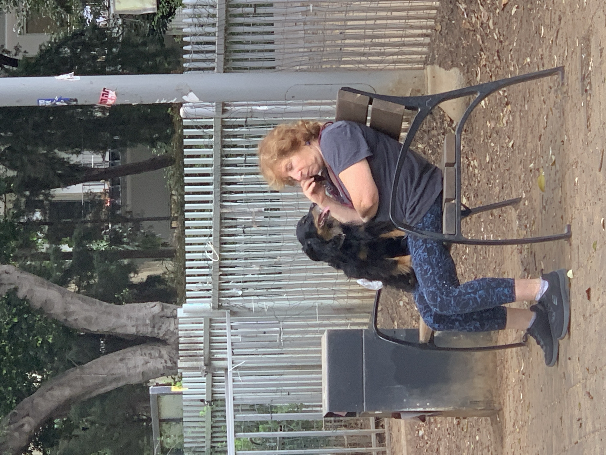 A woman with short auburn hair sits on a black metal park bench, talking on a mobile phone, with her dog standing between her legs, in a park in Tel-Aviv. The woman is wearing a grey short-sleeved t-shirt, blue floral-patterned cropped leggings, and black trainers with white socks. Her red lanyard is visible around her neck. The dog is black with brown markings on its face and legs. The bench is set on a paved area with loose leaves. A fence, trees, and a building are visible in the background. The setting appears to be an urban public park.