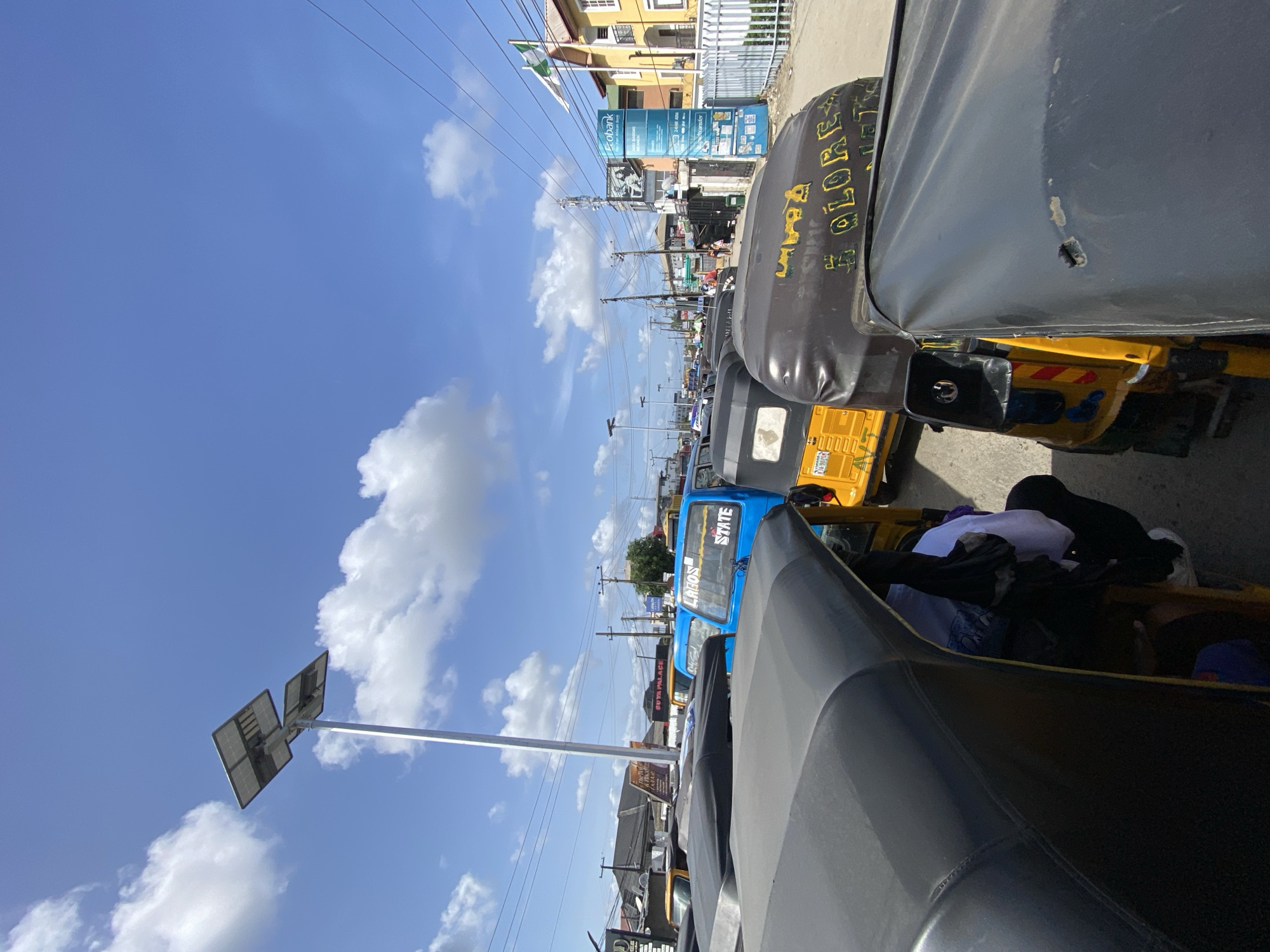The image shows a street scene in Ado, Nigeria. In the foreground are parked auto rickshaws ("keke"). A blue one in the middleground reads "LAGOS STATE". A solar street lamp stands to the left. In the background are buildings, signs, and people on the street. The Nigerian flag flies atop a yellow building. The sky is blue with scattered clouds.