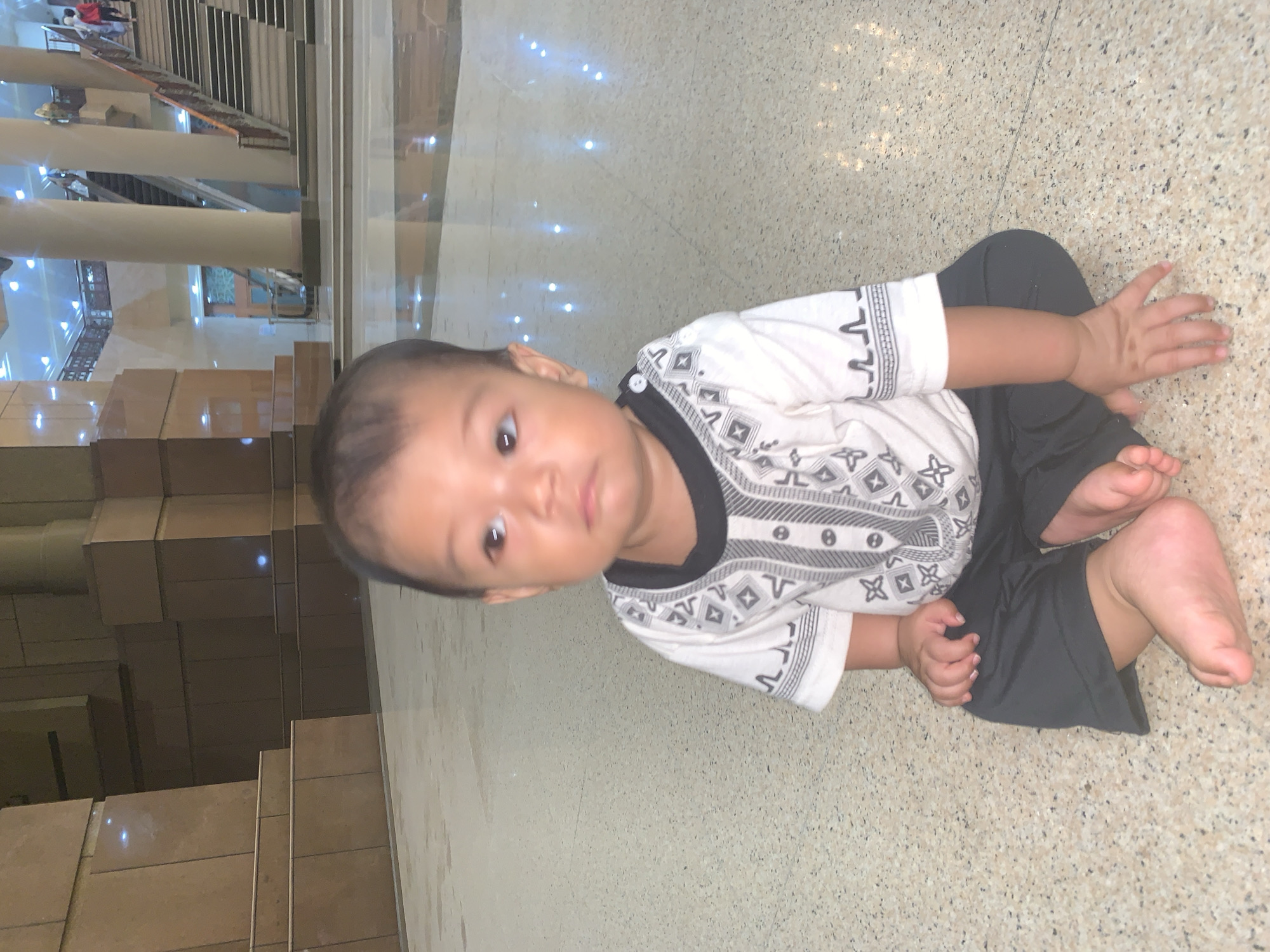 A young child, likely a baby or toddler, is the central subject, sitting on a light-colored, speckled tiled floor. The child wears a white short-sleeved shirt with black and grey geometric patterns and dark-colored pants. With dark hair and dark, inquisitive eyes, the child is looking directly towards the camera.
The setting is a spacious indoor public area, characterized by modern architecture. In the background, tall, light-colored pillars rise, and several flights of stairs or escalators are visible, suggesting a multi-level structure. Reflections of bright, artificial lights are scattered across the polished floor and other surfaces, indicating a well-lit interior. Distant, blurry figures of other people can be seen on upper levels. The environment appears to be a large public building, such as a shopping mall or a transport hub. No visible text is present. The scene takes place in Samarinda, Indonesia.