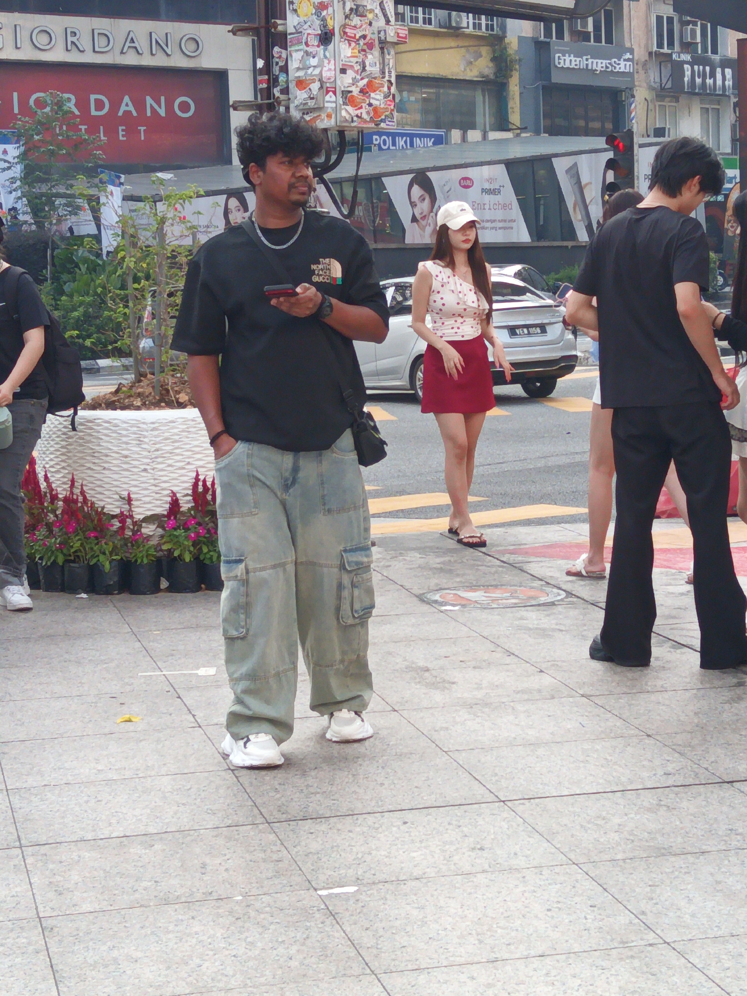 The image captures a street scene in Kuala Lumpur, Malaysia, likely along a commercial thoroughfare. In the foreground, a man with dark, curly hair stands facing left, holding a mobile phone in his right hand and wearing a black t-shirt with a "The North Face" logo, light-wash baggy cargo pants, and white sneakers. To his left, a person in a black t-shirt and dark pants is partially visible.
Further back, a woman in a white and red polka-dot sleeveless top and a short red skirt stands facing right, wearing a white baseball cap and flip-flops. She is standing near a silver car with a license plate that reads "YEN 1158". Behind the car, the exterior of several buildings is visible, including signage for "GIORDANO OUTLET", "Golden Fingers Salon", and "KLINIK". The environment includes a sidewalk with grey square tiles, potted plants with red flowers, and a large white planter. The background suggests a busy urban setting with traffic and commercial establishments.