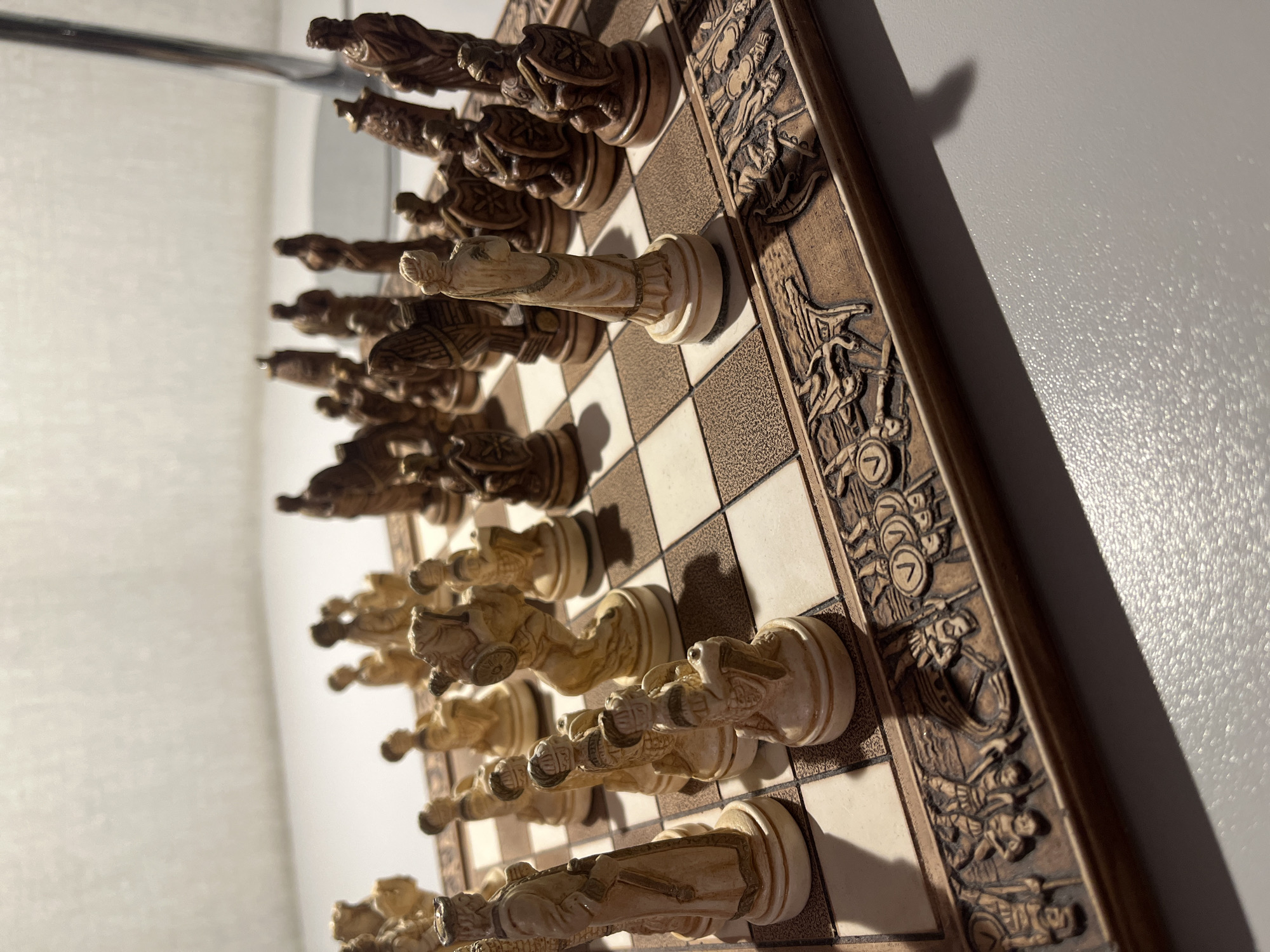 A close-up, eye-level shot shows a detailed, carved wooden chess set on a wooden chessboard. The pieces are in various stages of play. The light source appears to be from the upper right, casting shadows across the board. The background is out of focus, showing a white surface and a dark vertical element, possibly a lamp. The scene is set indoors on a flat, light-colored surface. There are no people visible in the image. The chessboard has an ornate carved border depicting figures and symbols. The chess pieces themselves are intricately carved, with some appearing to be in a lighter, aged ivory color and others in a darker wood tone. The pieces are arranged in a way that suggests an ongoing game, with some pieces advanced and others still in their starting positions.