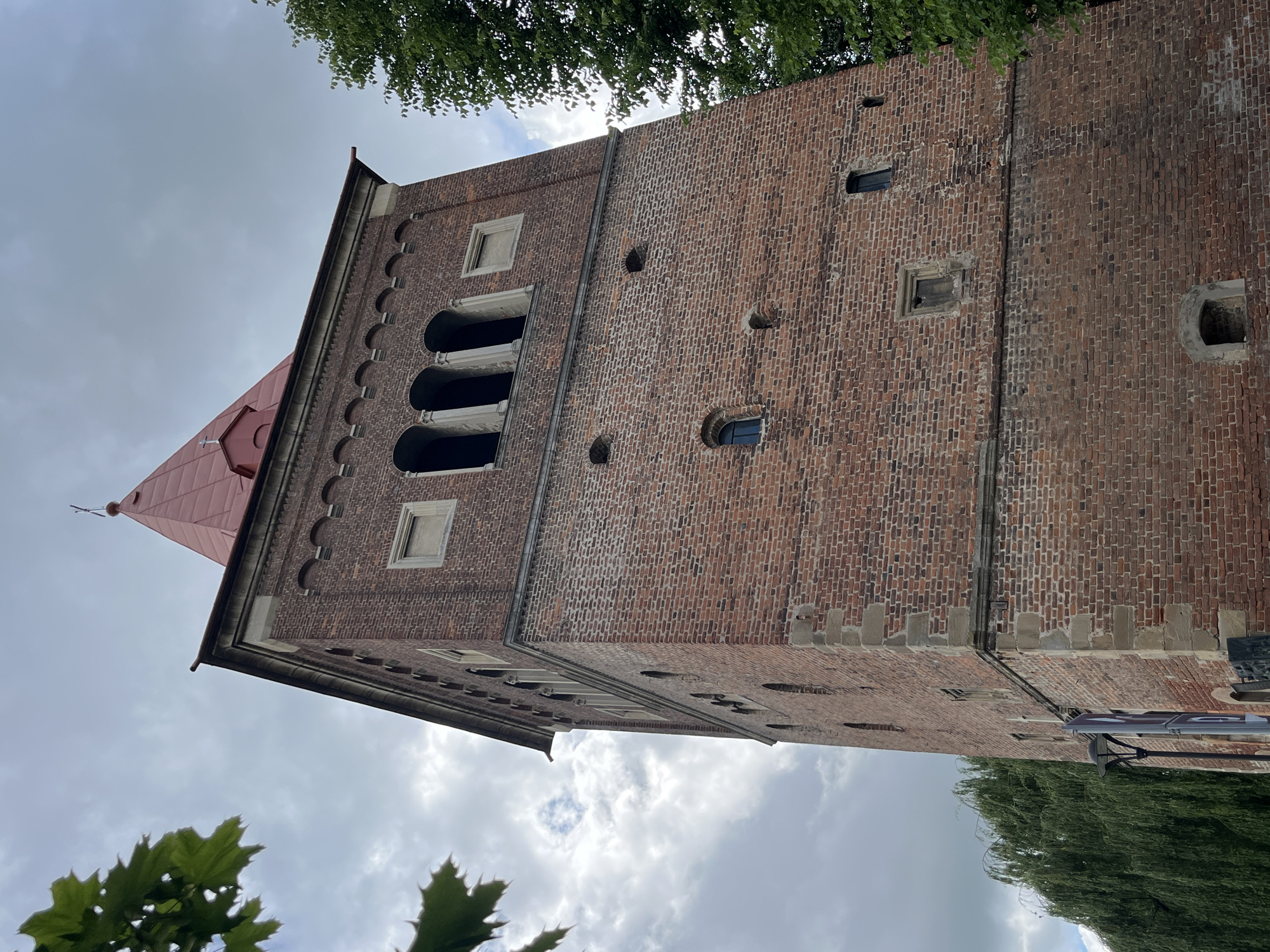 The image captures a close-up, low-angle view of the brick tower of the Church of Saint Bartholomew in Drohobych, Ukraine. The tower, constructed of weathered red brick, features several architectural elements, including arched openings near the top, small rectangular windows, and decorative stone accents at the corners. The roof of the tower is a steep, red, pyramidal structure topped with a small cross. Lush green foliage from trees frames the sides and top of the tower. The sky above is overcast with a mix of gray clouds and hints of blue. A lamppost is visible at the base of the tower on the left side of the image.