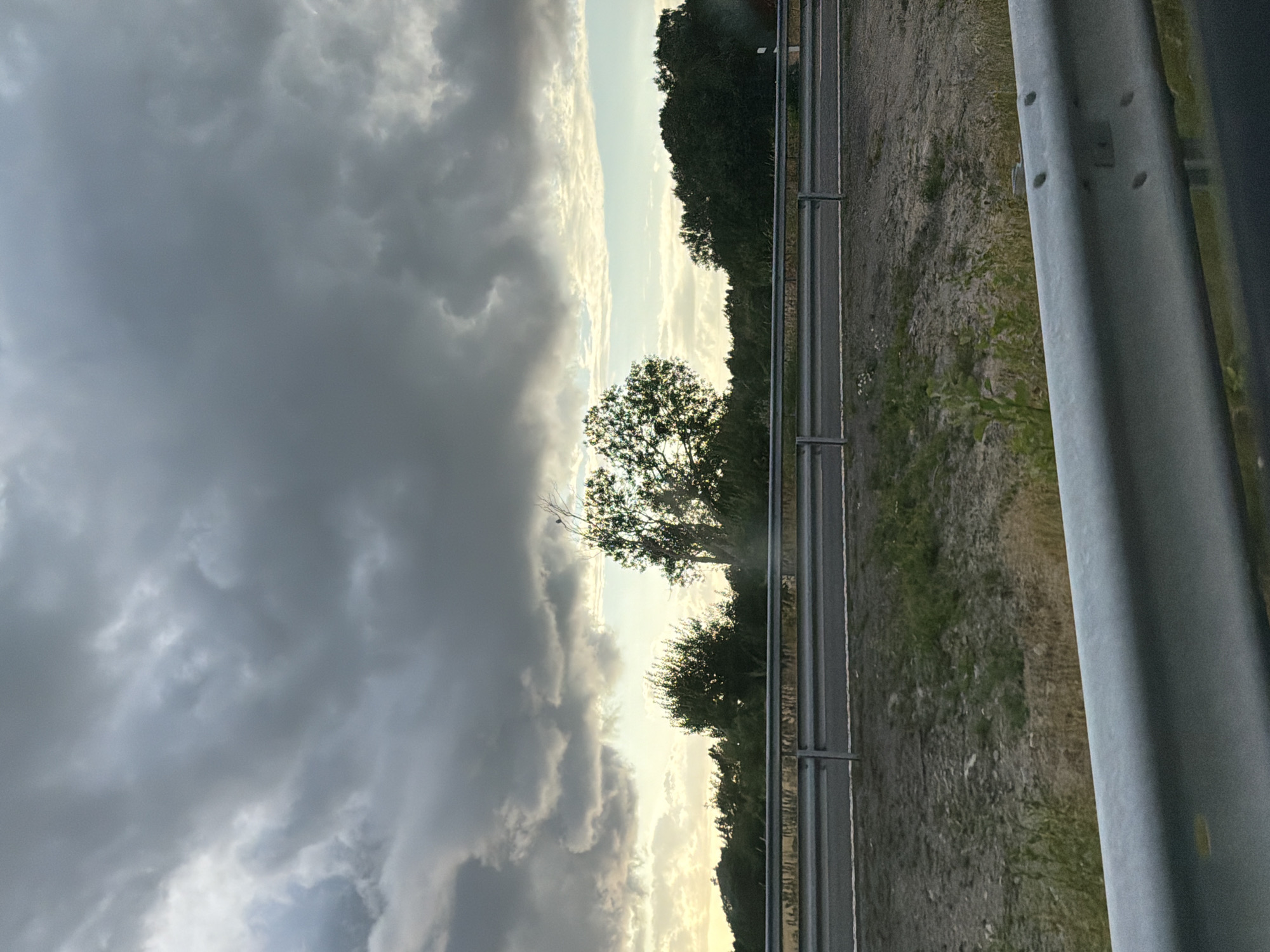 The image depicts a cloudy sky over a rural landscape. In the foreground, a metal guardrail is visible, suggesting the perspective of a vehicle on a road. Beyond the guardrail, a grey asphalt road is present, with a grassy verge and some sparse vegetation. A second, lower guardrail runs parallel to the road. Further in the background, a line of trees forms a dense natural barrier, with a few taller trees interspersed. The sky is dominated by dark, heavy clouds, with patches of lighter sky and some hints of sunlight breaking through near the horizon. The scene appears to be located in Limabacka, Sweden. There are no people or animals visible in the image.