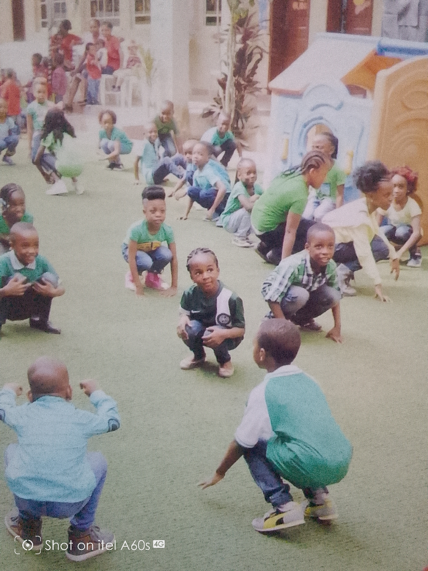 A group of young children are outdoors on a green artificial surface. They are mostly squatting or crouching. Most children wear green or blue shirts and jeans or trousers. In the background are indistinct people and a light-colored play structure. The location is Idimu, Nigeria, as implied by the camera watermark.
