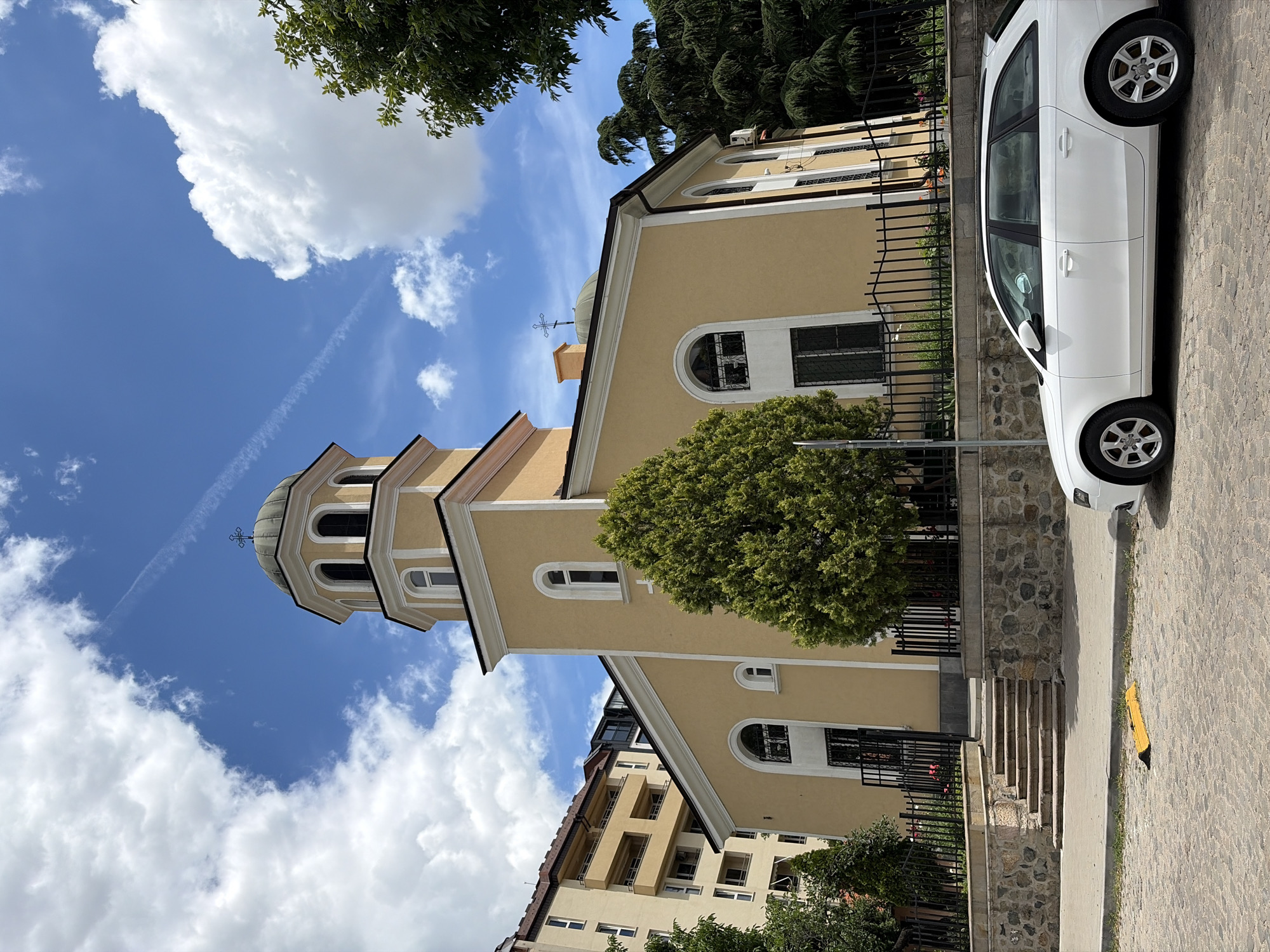 The image shows the exterior of the St. Nicholas the Miracle-Worker Church in Sofia, Bulgaria. The church has a yellow facade, a bell tower with a dome, and a cross on top. A white Audi station wagon is parked in front of the church. The sky is blue with white clouds. There are trees and a modern building in the background. The ground in front of the church is paved with cobblestones.