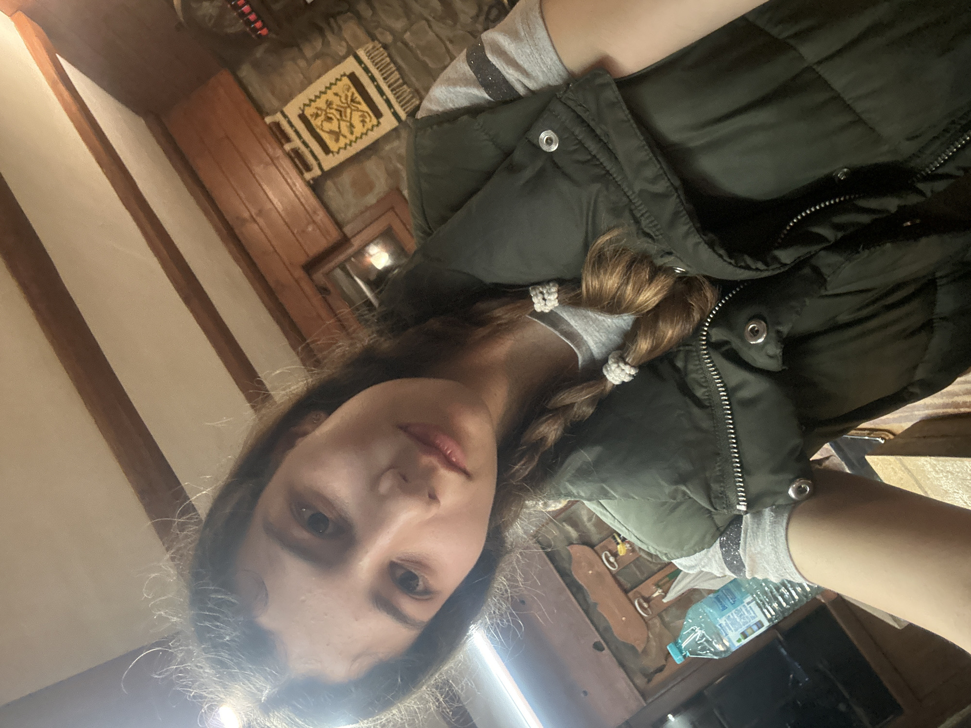 A sideways-oriented selfie captures a young woman indoors in Ignatovtsi, Bulgaria. The woman, with medium-length brown hair styled in two braids secured by white, possibly beaded, ties, looks directly at the camera with a neutral expression. She is dressed in a light grey long-sleeve shirt with subtle sparkly details on the sleeves, worn under a dark green puffer vest.
The setting is a rustic interior featuring a textured stone wall in the background, accented by a small, decorative woven tapestry or rug displaying a yellow and brown geometric pattern. Prominent wooden beams are visible on the ceiling or upper wall, contributing to the traditional aesthetic. A plastic water bottle with a blue cap and an unreadable label is visible to the lower right of the frame, resting on a surface. The indoor lighting suggests the time of day is not bright daylight, possibly evening or an overcast day. No specific activity other than taking the selfie is evident.