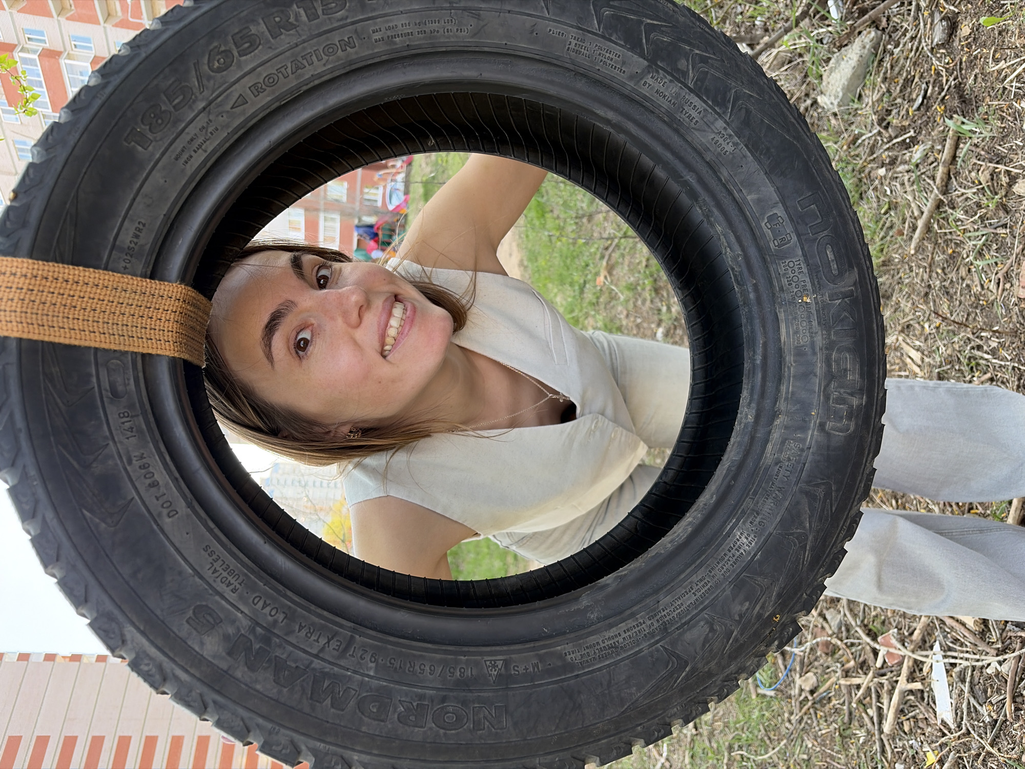 The scene depicts a young woman playfully positioned inside a tire swing. The tire, identified as a "NORDMAN 5" model with size "185/65R15 92T," is suspended by a tan strap. The woman is smiling and looking at the camera, wearing a light-colored vest or top and light-wash denim pants. The background shows hints of an urban environment, including a building with visible windows and some green vegetation. The tire's markings indicate it was made in Russia by Nokian Tyres. While the OCR mentions "49005", that number could be referencing its location in Izhevsk, Russia.