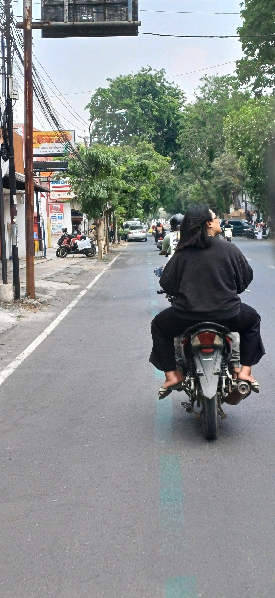 The image shows a street scene in Kecamatan Pati, Indonesia. A person wearing a black top and black pants is riding a motorcycle. The motorcycle is dark in color with some silver accents. The rider has long, dark hair and is wearing glasses. They are on a paved road with a white line on the left side, indicating a lane marker or the edge of the road.
On the left side of the road, there are buildings and shops, with some of them having signage in Indonesian. One sign displays "KAB. PATI", indicating the administrative region. There are also utility poles with numerous wires. Trees line the right side of the road, and a few other motorcycles and a white car are visible further down the road. The sky is overcast, suggesting cloudy weather. The overall impression is a typical street scene in a populated area of Indonesia.
