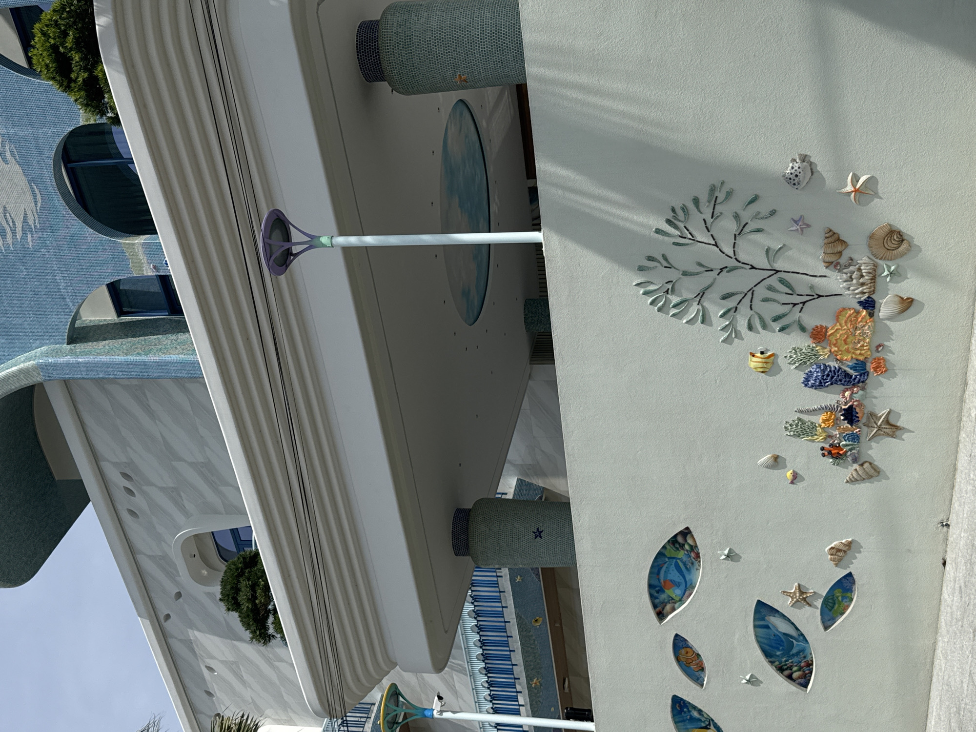 The image shows the exterior of a building with a distinctive ocean-themed design. The building features light blue mosaic tiles with wave patterns and arched windows. In the foreground, there is a light mint green wall decorated with colorful ceramic art depicting marine life, including fish, coral, starfish, and seashells. There are also several oval-shaped openings in the wall, revealing underwater scenes with aquatic creatures. The scene is set in a city environment, likely a resort or public space, given the artistic embellishments. The lighting suggests it is daytime, with shadows cast by palm trees. The overall impression is of a vibrant and whimsical architectural design that celebrates the ocean.