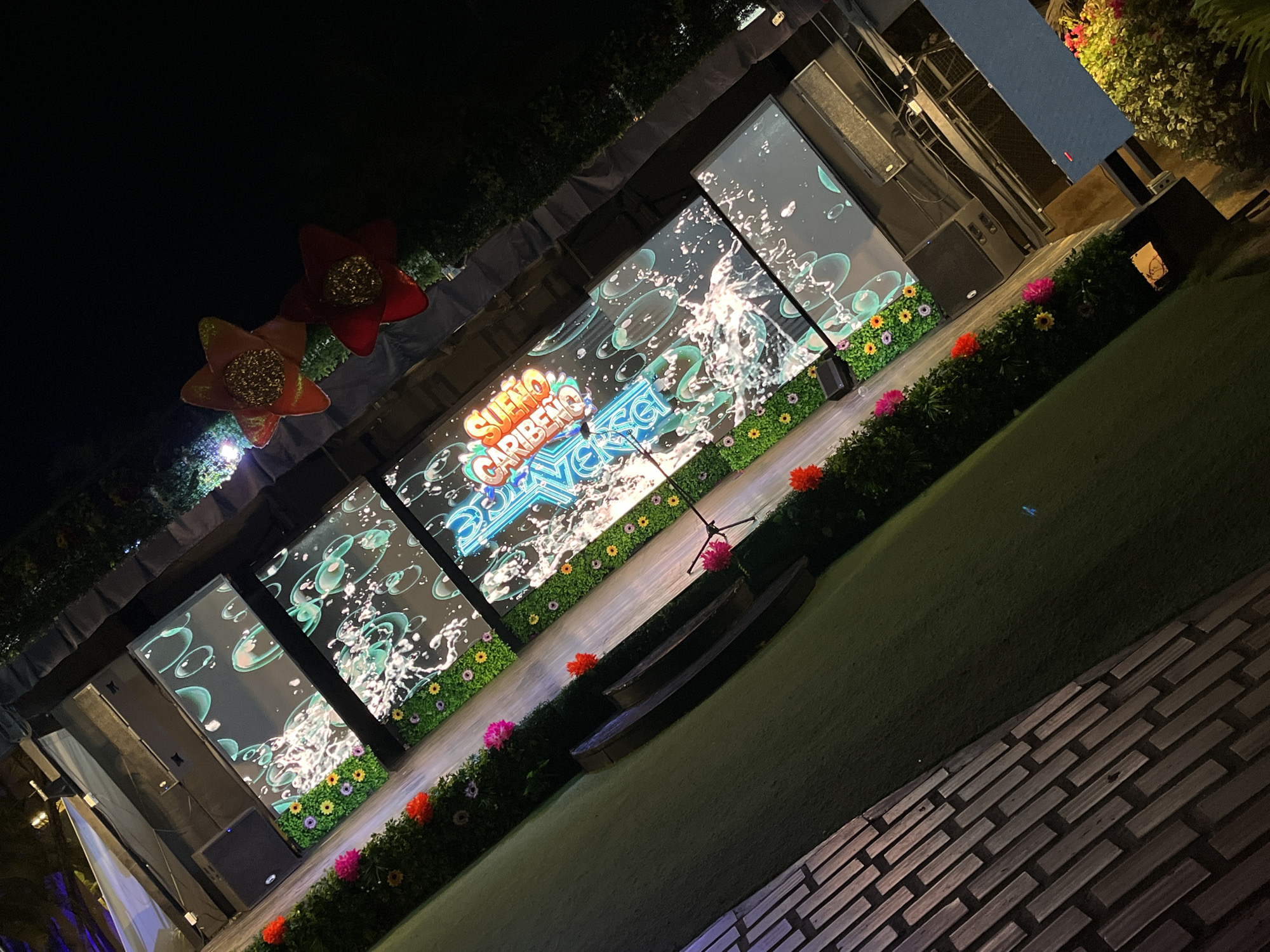 The image depicts an outdoor stage in Porlamar, Venezuela, at night. The stage is decorated with floral arrangements and features three large video screens displaying a "Sueno Caribeno" (Caribbean Dream) theme with water and bubble imagery. Two decorative flower-shaped lights adorn the top left corner of the stage. In front of the stage, there is a grassy area with a wooden bench and a brick-paved walkway. There are no visible people in the frame. The overall atmosphere suggests an event or performance venue.