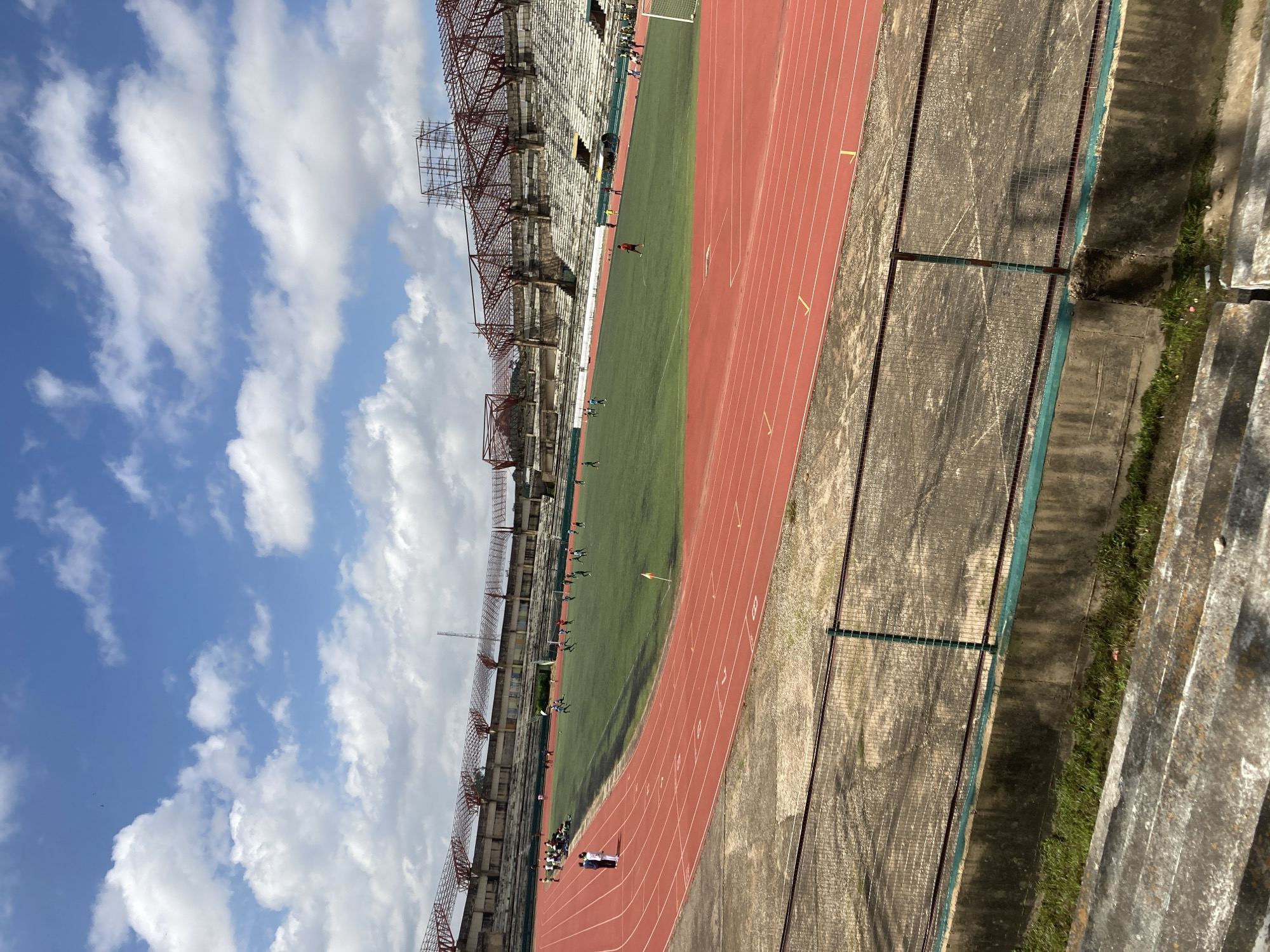 The image depicts a wide shot of the newly constructed National Stadium in Abuja, Nigeria. The stadium features a large, green football field surrounded by a red athletic track. Several individuals, appearing to be athletes, are present on the field, some engaged in what looks like a football practice. The stands of the stadium are visible, with a predominantly empty seating area. The architecture of the stadium is modern, with exposed metal structures supporting the roof. The sky above is a bright blue with scattered white clouds. The foreground shows parts of the stadium's lower seating area, which appears to be made of concrete and has some greenery growing around it. The overall scene suggests a sports facility in use on a clear day.