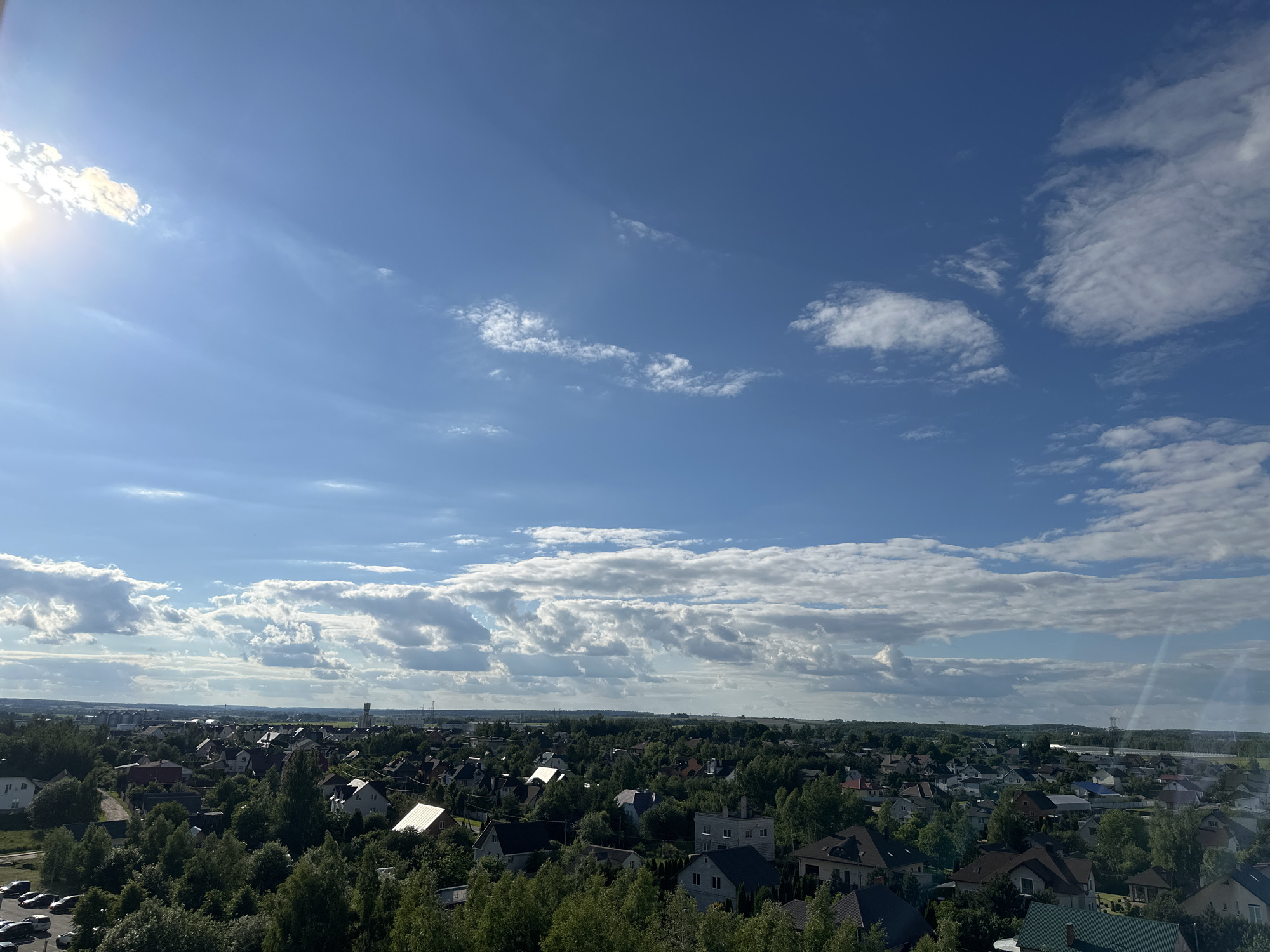 This is an aerial, wide-angle photograph of a suburban residential area bathed in bright sunlight under a blue sky with scattered clouds. The scene depicts numerous houses with pitched roofs, interspersed with abundant green trees. The buildings vary in color and architectural style, with some appearing to be multi-story. In the lower left corner, a parking lot with several cars is visible. The horizon line is low in the frame, emphasizing the expansive sky. The location is identified as the Shchomyslicki Sel'ski Savet in Belarus. The overall impression is of a tranquil, sun-drenched day in a well-established residential community.