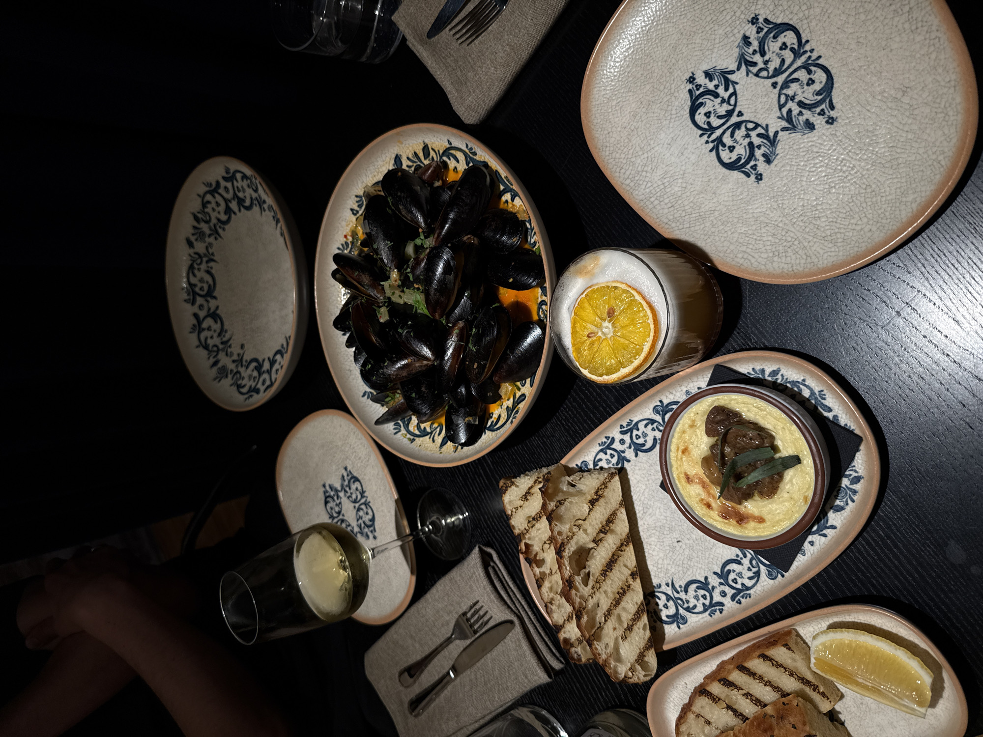 A dimly lit dining table is captured from a high angle, likely depicting a restaurant scene in Riga, Latvia, during the evening or night. The table is set with various dishes and drinks, showcasing an elegant yet rustic aesthetic.
At the center, a large oval platter holds a generous portion of mussels in a vibrant orange-yellow sauce, garnished with fresh green herbs. Surrounding it are several decorative ceramic plates with a distinct cracked glaze texture and intricate blue floral patterns. Two round plates are visible, one on the left and another partially on the upper right. An additional oval plate on the lower right features a small, round bowl containing a creamy dip or pâté, topped with dark pieces of meat or mushrooms and a few green sage leaves. Accompanying this dish are several slices of grilled bread, showing prominent char marks, and a fresh lemon wedge on a smaller plate.
Beverages include a stemmed glass holding white wine, situated near the partially visible arm of a diner on the lower left. Another tall glass, positioned on the upper right, contains a frothy drink garnished with a bright orange slice. A neatly folded grey linen napkin with a fork and knife set rests on the table. The table surface itself is dark, possibly a dark wood, reflecting some ambient light. No discernible text is visible within the image.