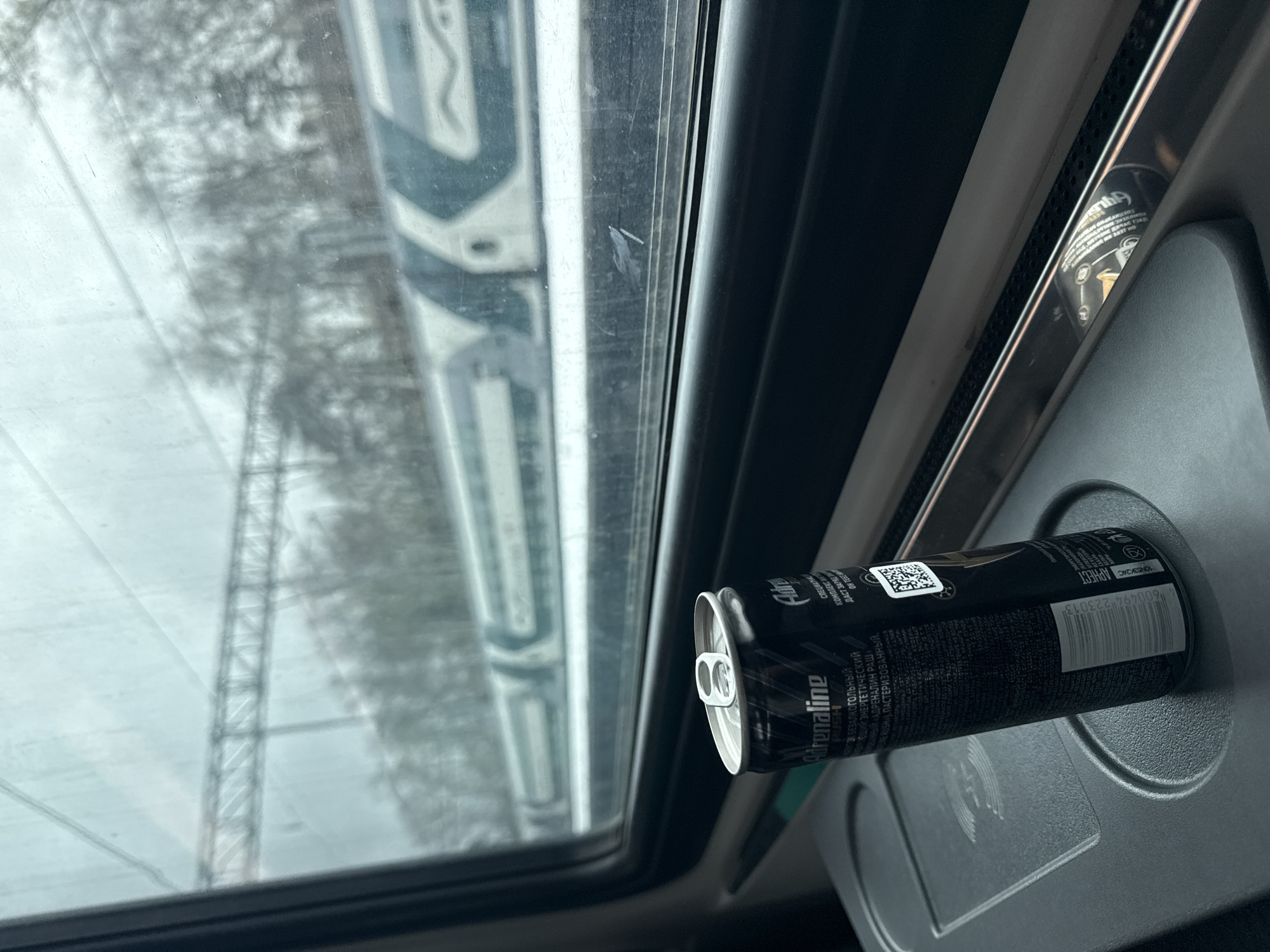 The image depicts an indoor scene, likely within a train traveling through Mytishchi, Russia. A black energy drink can, "Adrenaline Rush," is centrally featured, resting horizontally in a built-in cup holder or recess in the vehicle's dark gray interior paneling. The can's silver pop-top is visible on the left.
Through the window, the exterior appears blurry, indicating the train is in motion. The landscape outside is overcast and seemingly cold, with bare, indistinct trees and a faint power line tower visible. Another train or large structure is partially visible through the window's reflection or transparency, further confirming the train setting. The window itself shows some streaks or dirt.
No specific activity is taking place, but the presence of the energy drink suggests someone is traveling. The time of day appears to be daytime, under cloudy or gloomy weather conditions.
Visible text on the can, primarily in Cyrillic, includes "Adrenaline RUSH," "БЕЗАЛКОГОЛЬНЫЙ" (non-alcoholic), "ЭНЕРГЕТИЧЕСКИЙ" (energetic), and "ПАСТЕРИЗОВАННЫЙ" (pasteurized). Phrases like "ДАСТ ЗАРЯД" (will give energy/charge) and "ОН ТЕБЕ" (for you) are also legible. A QR code and a barcode with the number "600494 223013" are clearly displayed on the can's label.