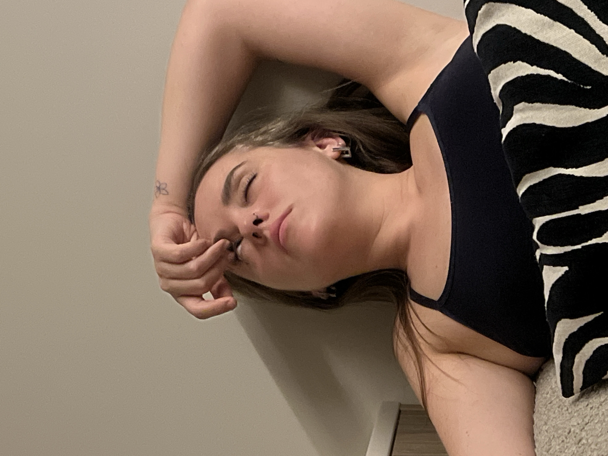 A woman is lying down, eyes closed, with her right arm raised above her head. She is wearing a dark-colored tank top and has a nose piercing. A black and white zebra-striped object is next to her. There is a tattoo on her wrist. The background consists of a light-colored wall. The location is not discernible from the image.
