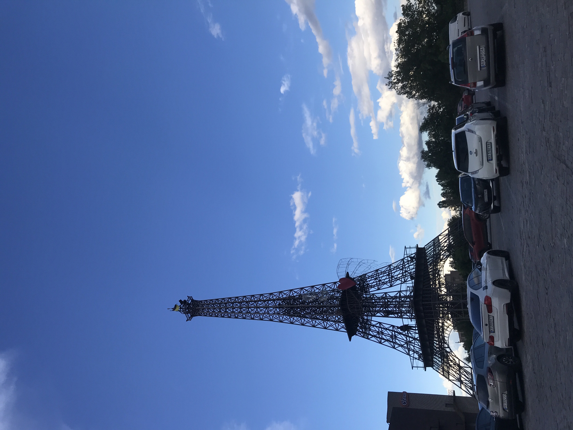 The image shows a replica of the Eiffel Tower located in Kharkiv, Ukraine. It is a sunny day with a clear blue sky and some scattered clouds. The Eiffel Tower replica is made of metal and stands tall, with a decorative heart-shaped structure on its midsection. At the base of the tower, there is a parking lot with several parked cars. The car license plates visible are Ukrainian. A building with a “Celentano” sign is near the tower. Trees are in the background.