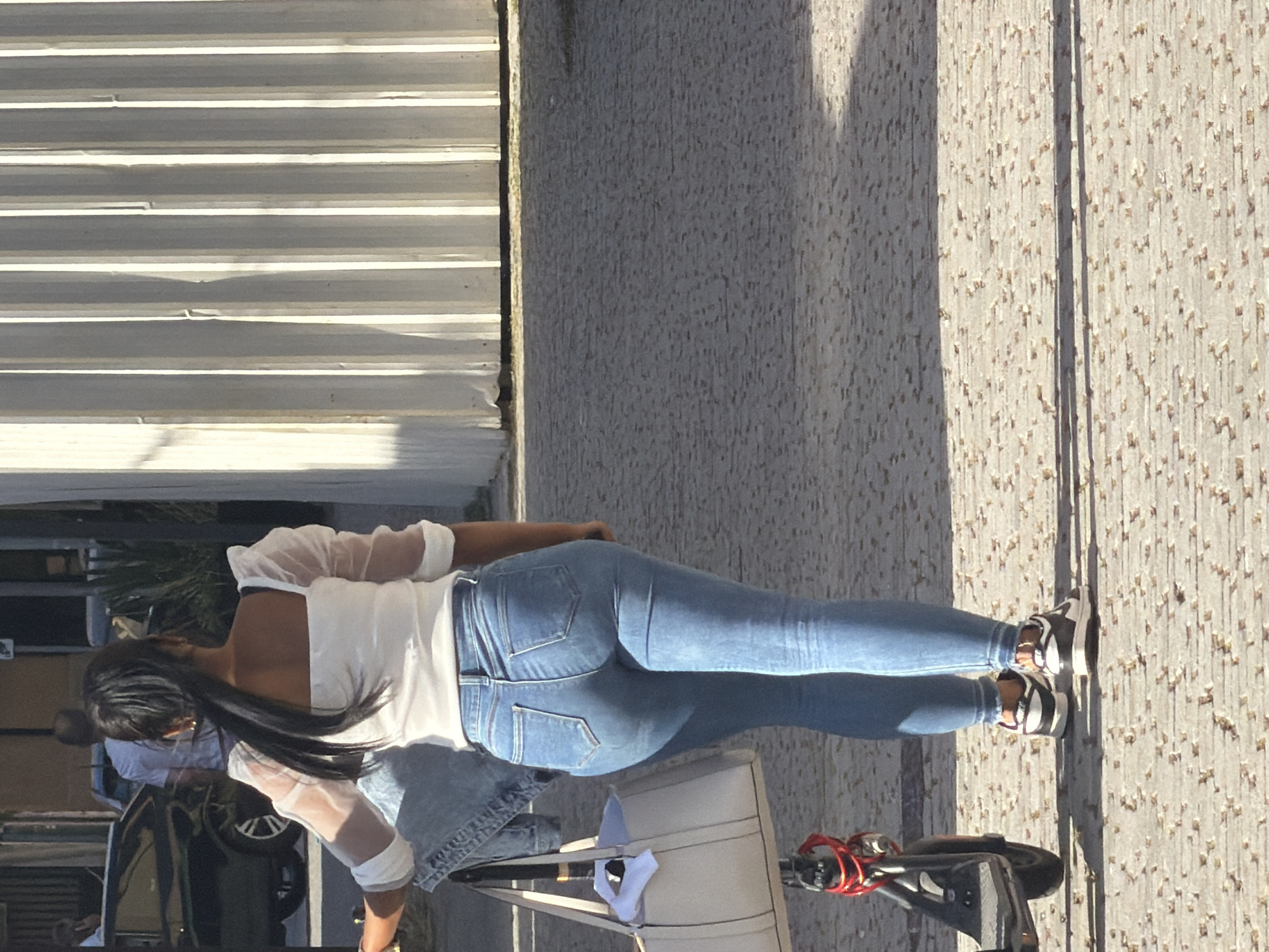 A woman with her back to the camera, wearing a white sheer-sleeved top and fitted blue jeans, walks on a paved walkway in Espinho, Portugal. She has her dark hair pulled back in a ponytail. She is carrying a large light-colored tote bag on her left arm, and a denim jacket is draped over her shoulder. A black electric scooter is parked next to her on the left. The background features a corrugated metal wall and the shadow of the woman and scooter on the ground.