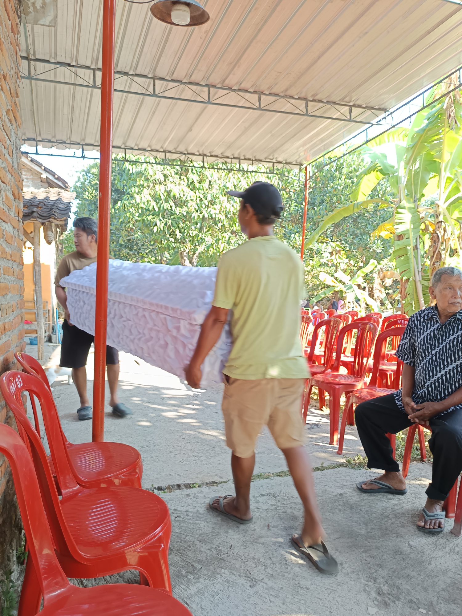 In Geneng, Indonesia, two men carry a white coffin covered in fabric. One man, wearing a light green t-shirt and tan shorts, supports the coffin from the right side, while another man in a brown t-shirt and black shorts carries it from the left. They are walking on a concrete path under a covered structure with a corrugated metal roof. Red plastic chairs are arranged in rows to the right of the path, and an older man in a patterned shirt and black pants is seated on one of these chairs. A brick wall is visible on the left side of the image, and lush green foliage and banana trees are present in the background, indicating an outdoor setting. The scene appears to be a funeral or a related ceremony.