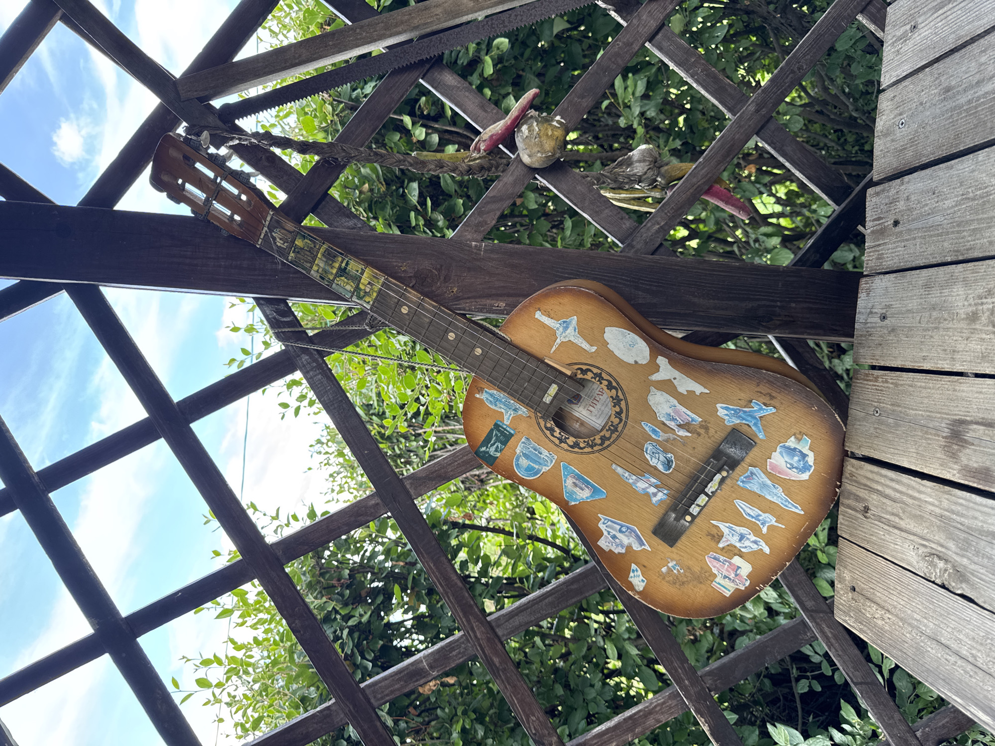 A brown acoustic guitar adorned with various stickers is propped against a dark wooden lattice structure in an outdoor setting. The lattice forms a diamond pattern, with green foliage visible through the openings. A wooden plank runs across the bottom foreground. The sky, visible through the lattice, is blue with scattered clouds. A saw and a rope with dried items hanging from it are also attached to the lattice. The guitar has a worn appearance and features a circular soundhole with intricate detailing. The neck of the guitar has colorful stickers applied to it. The overall scene suggests an outdoor, possibly rustic, environment. The location is specified as Gubarevo, Russia. There are no people present in the image.