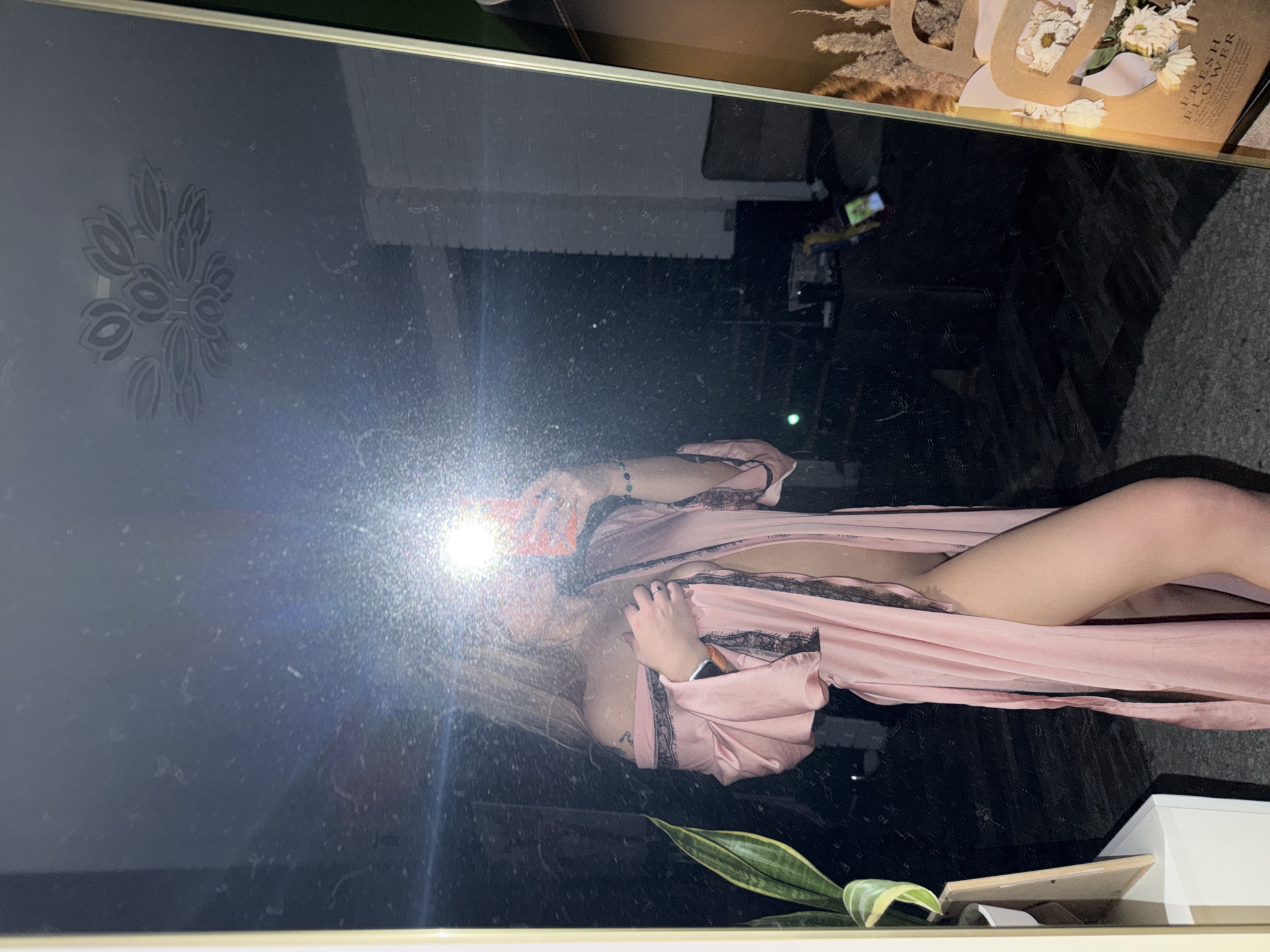 A reflection of a woman in a mirror. The woman is wearing a pink robe with black lace trim. The reflection shows a portion of her body and a hint of undergarments. The surrounding environment includes a plant in a pot, a box with "FRESH FLOWER" written on it, and a wall with a decorative pattern. It is not possible to ascertain the exact location within Большая Шильна from the image.
